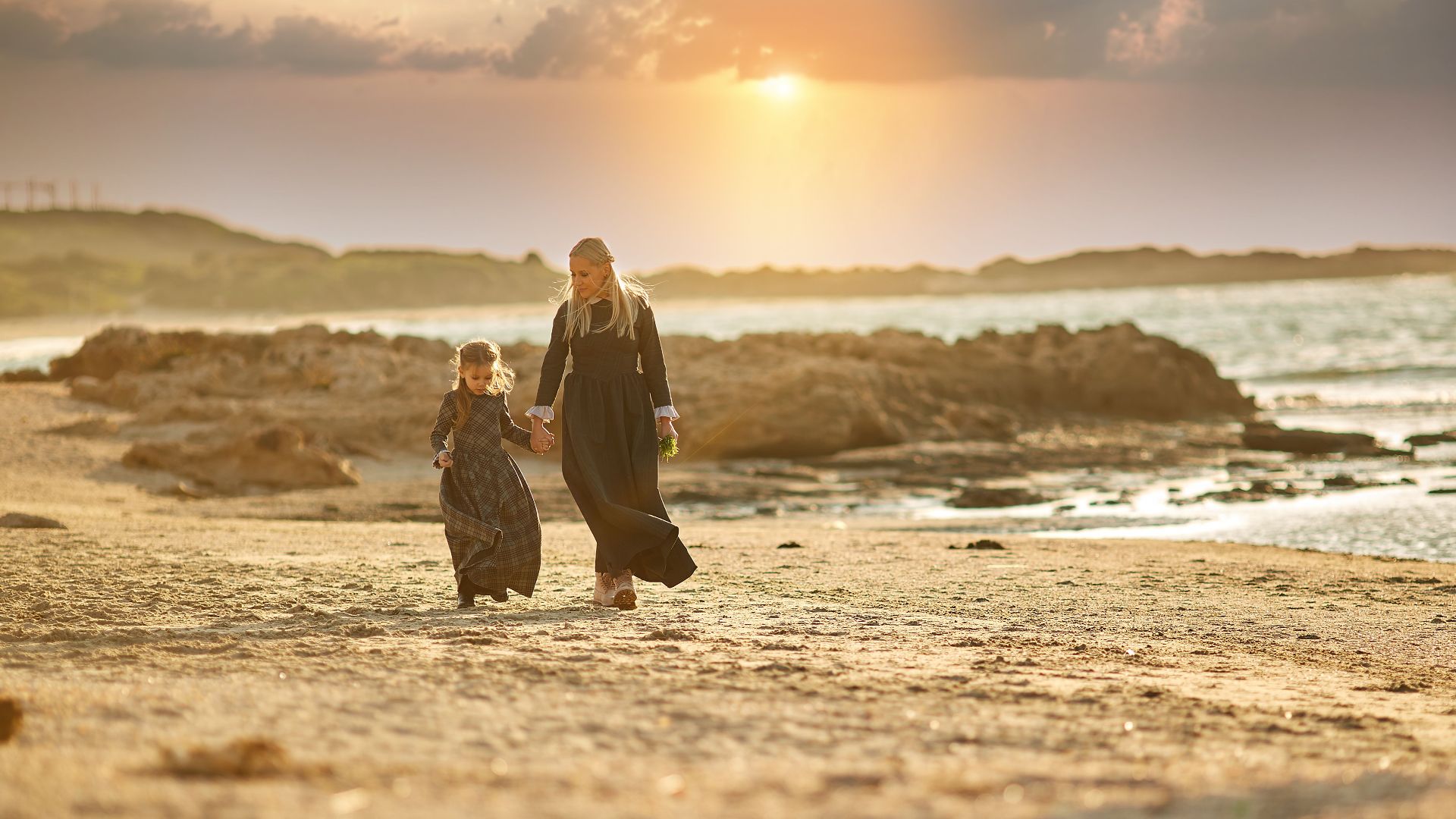 Mother and daughter walking hand in hand on a beach during a beautiful sunset, creating a serene family moment.