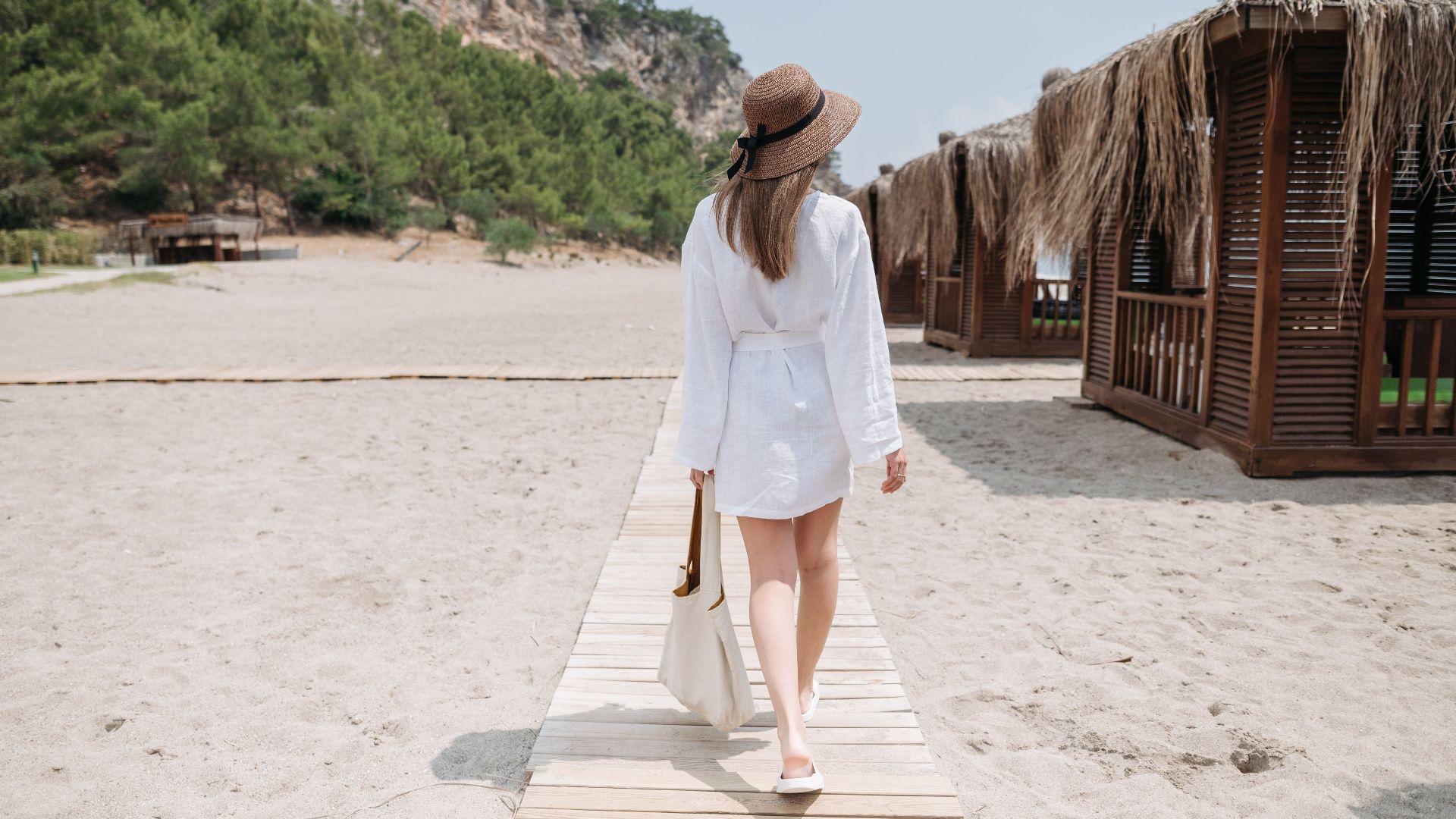 A woman in a white dress walks on a beach boardwalk surrounded by thatched cabanas, enjoying a summer day.