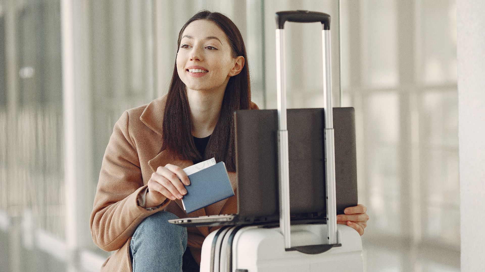 Smiling adult ethnic female traveler in trendy coat holding passport and using laptop on luggage in airport corridor for checking ticket for correctness