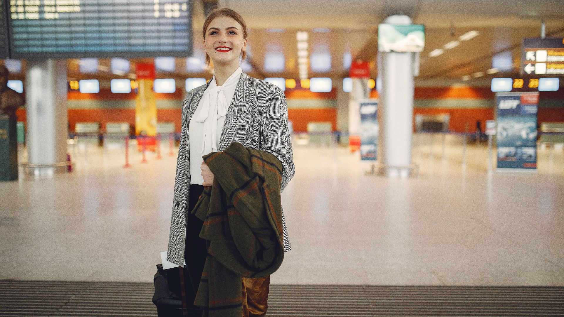 Smiling female manager in elegant wear checking schedule of flights in airport while standing with suitcase and coat looking up