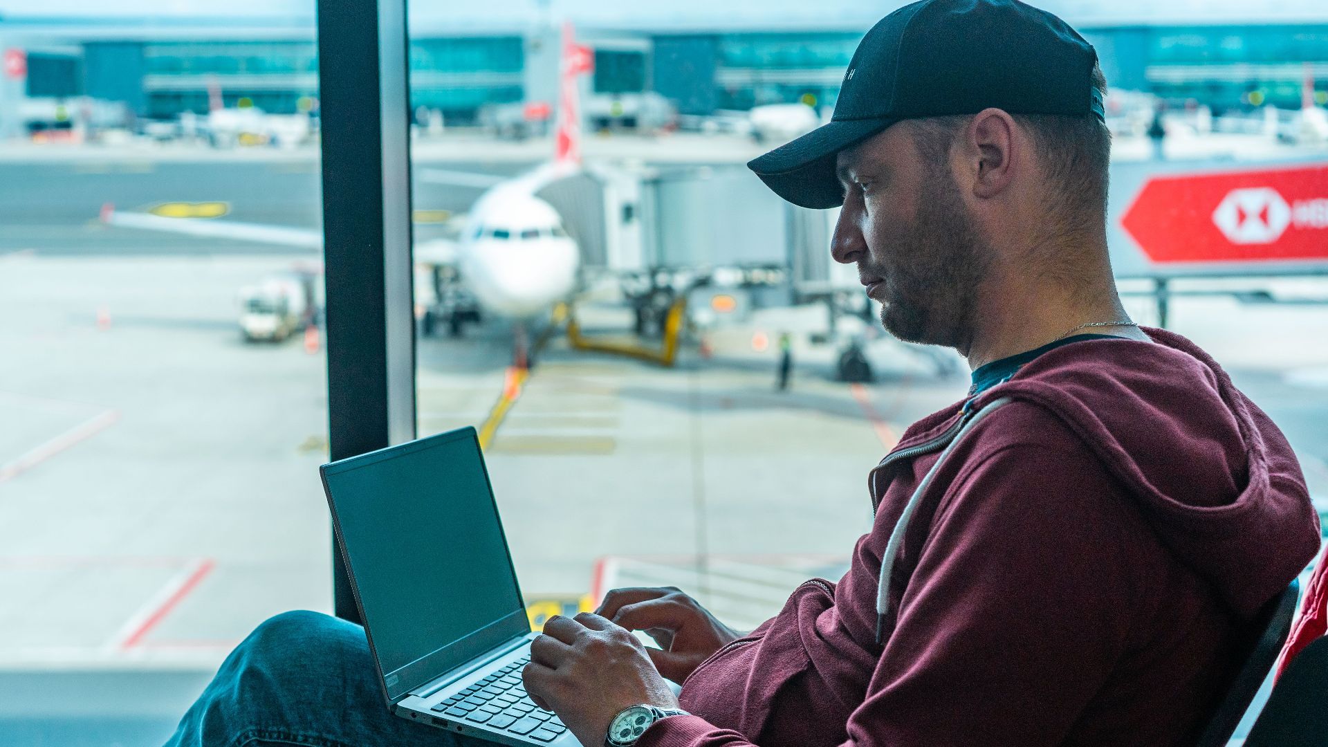 A man working on his laptop in an airport terminal, with a plane visible outside.