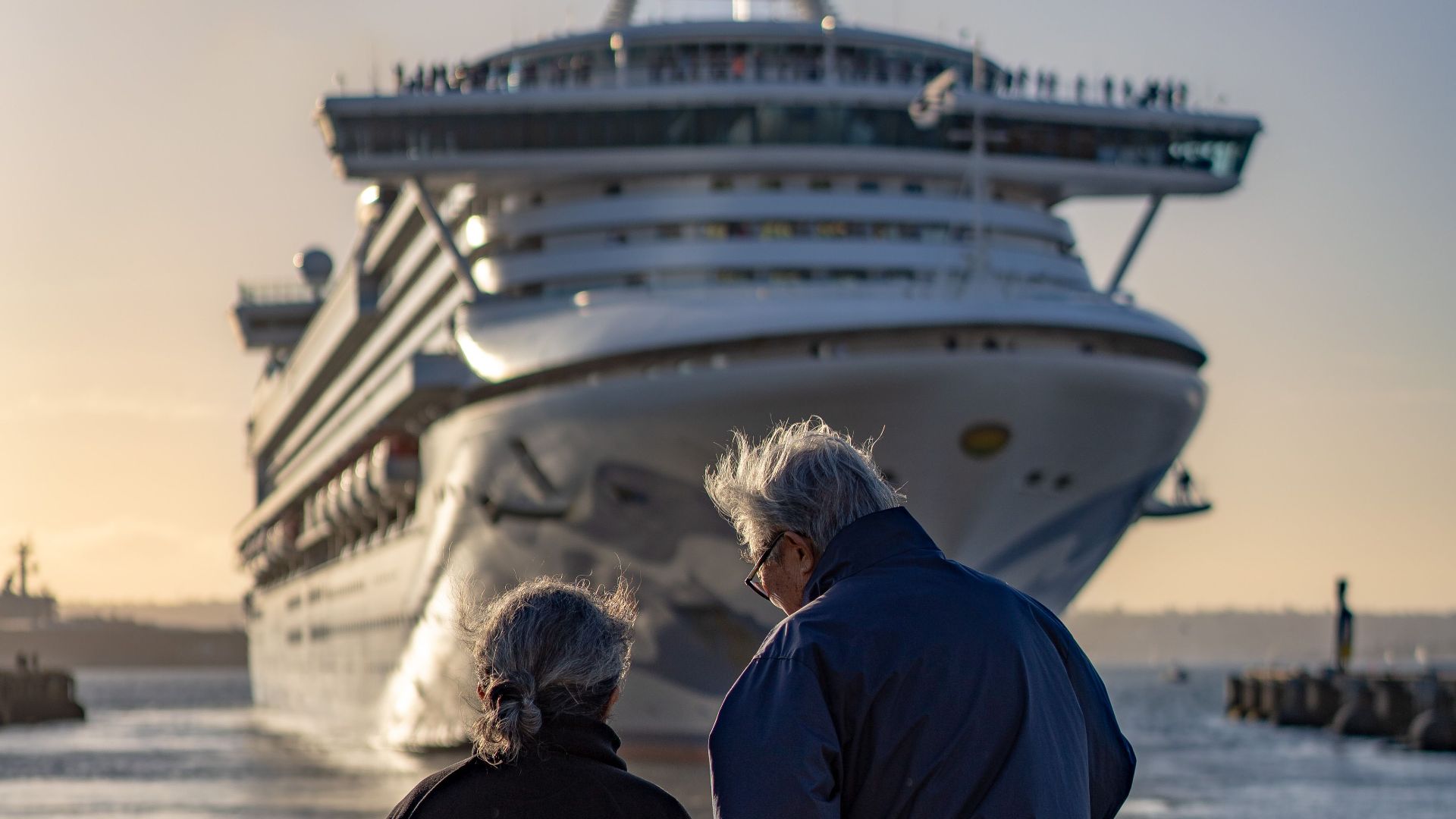 Senior couple standing at railing, admiring large cruise ship approaching dock at sunset.
