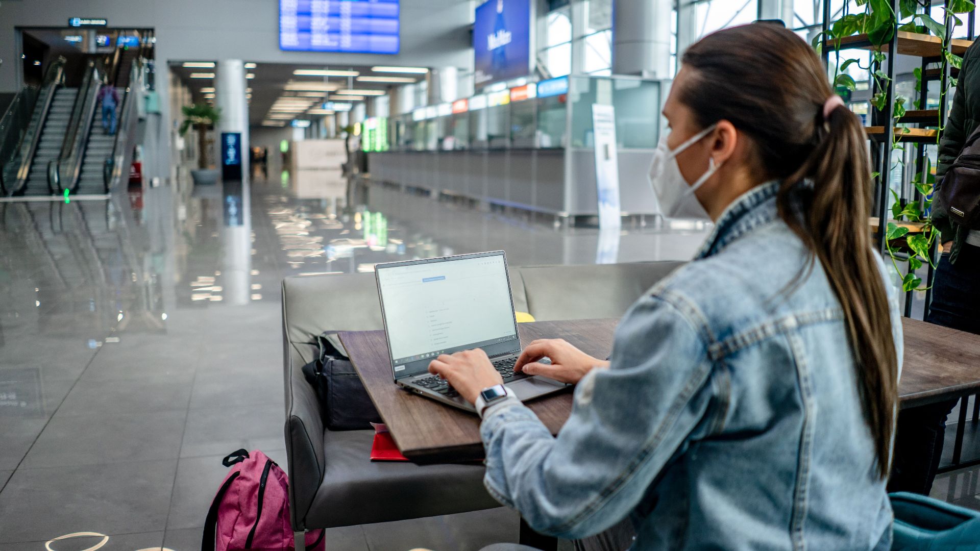 Side view of concentrated female freelancer in protective mask typing on netbook while sitting at table in modern airport terminal