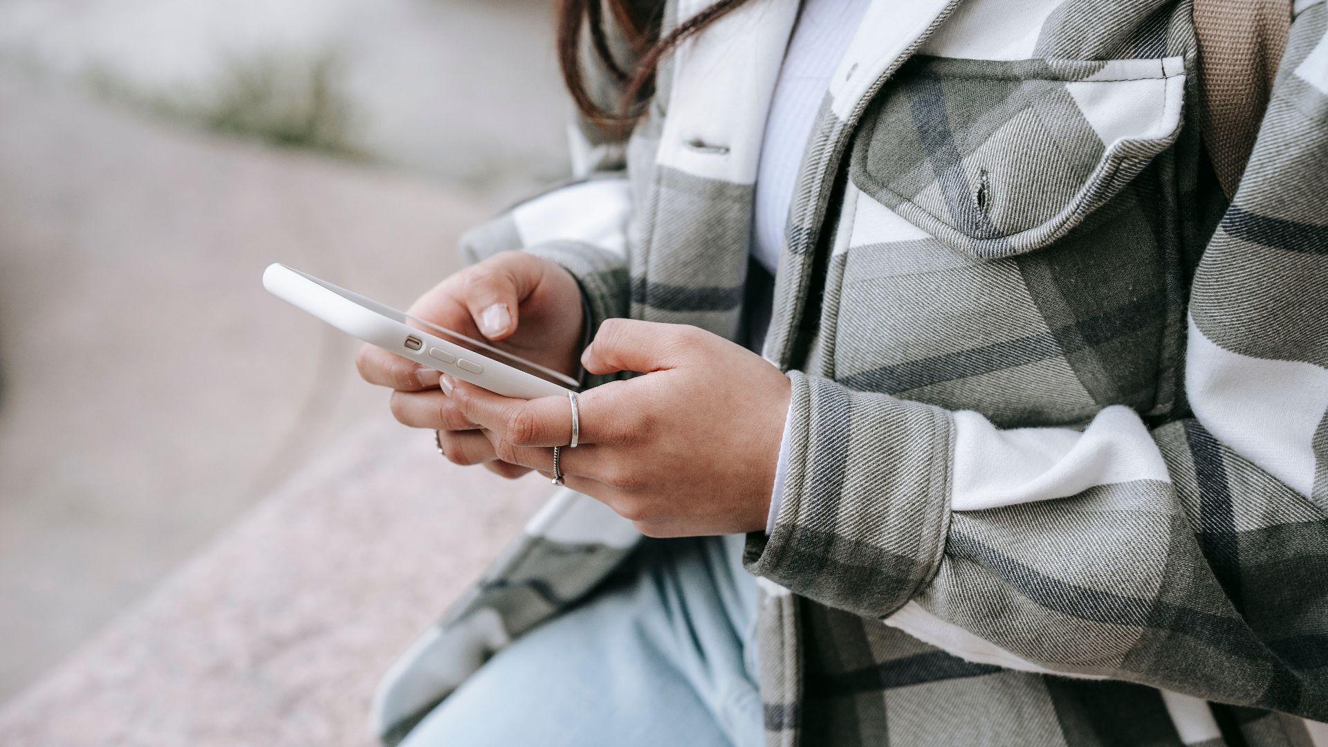 Crop anonymous young woman in casual outfit sitting on stone fence and text messaging on phone in daytime