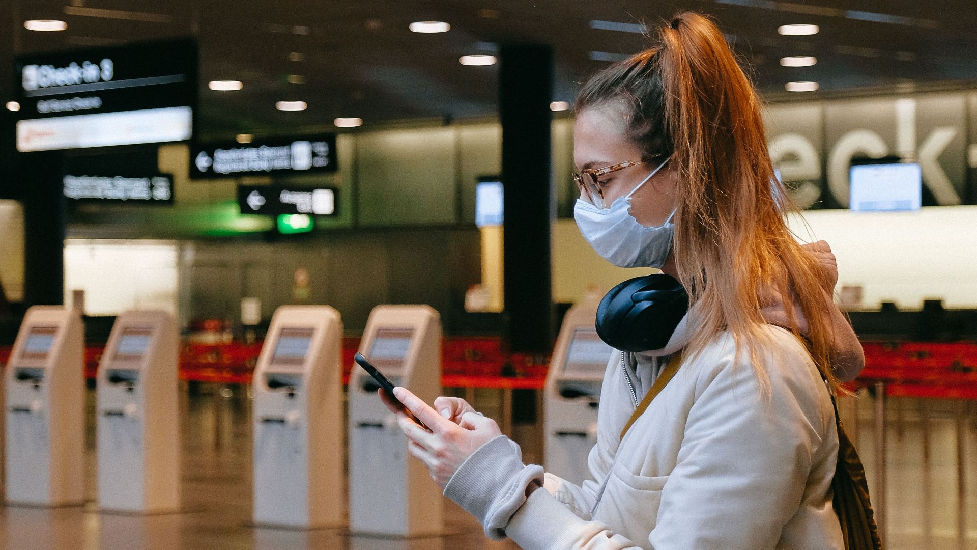 Woman wearing a mask, texting in airport terminal with signage in background.
