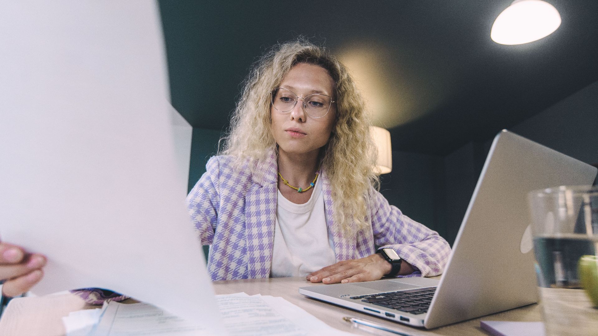Woman in an office environment reviewing documents with focus, surrounded by technology.