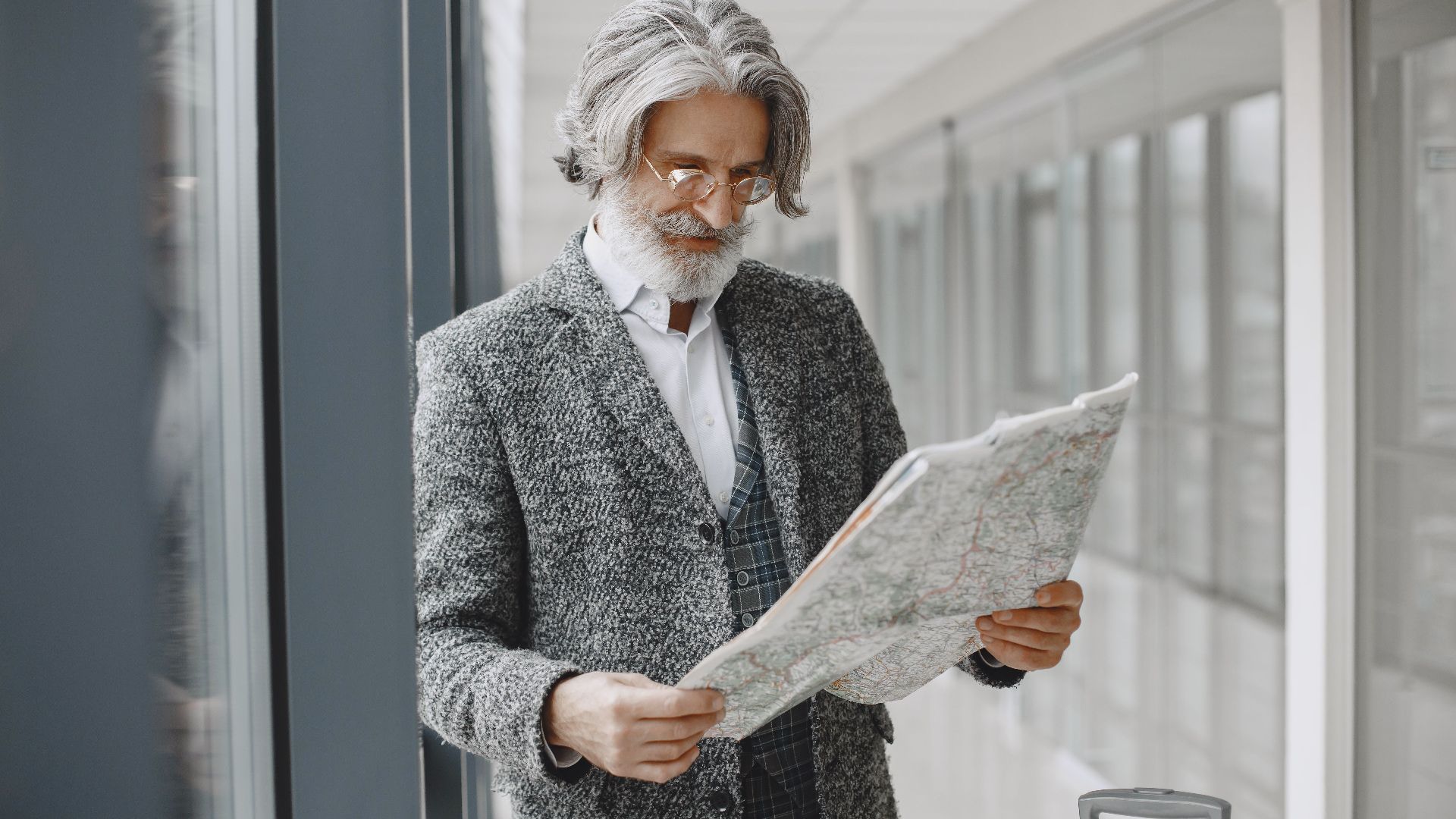 Elderly man with suitcase reads map in a bright airport corridor, planning his journey.