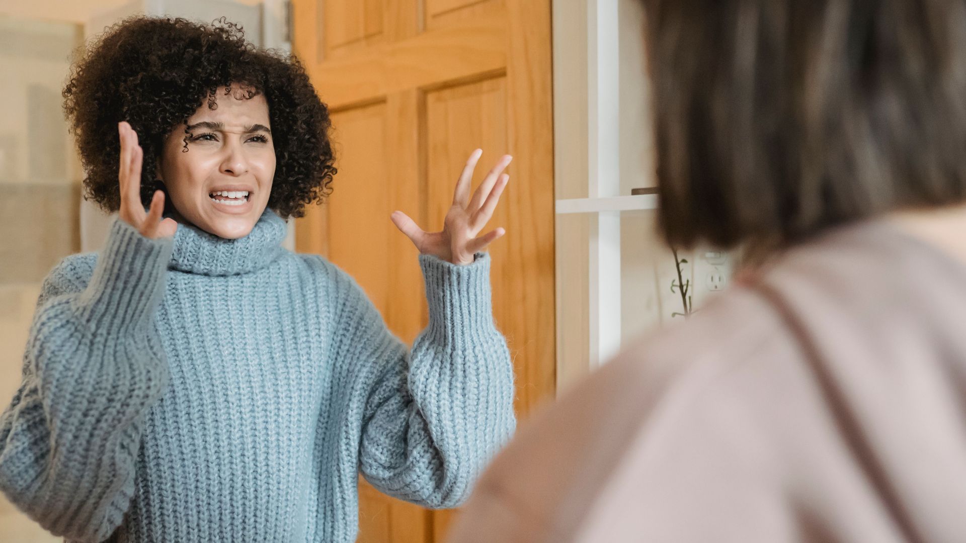 Angry young multiracial ladies in casual clothes standing in bright apartment near door while having disagreement and looking at each other