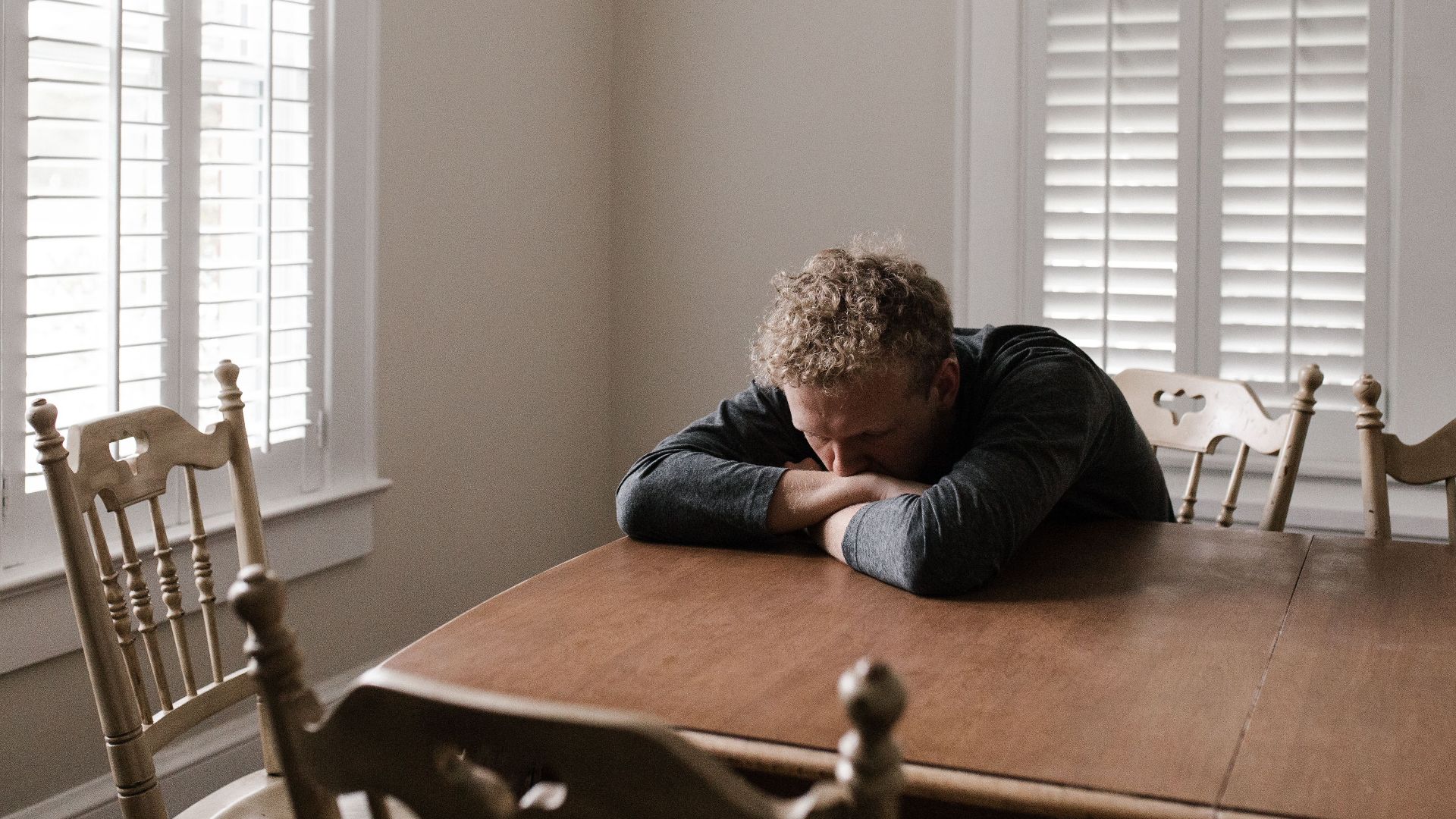 An adult man with emotions of sadness and anxiety sitting alone at a wooden table near windows.