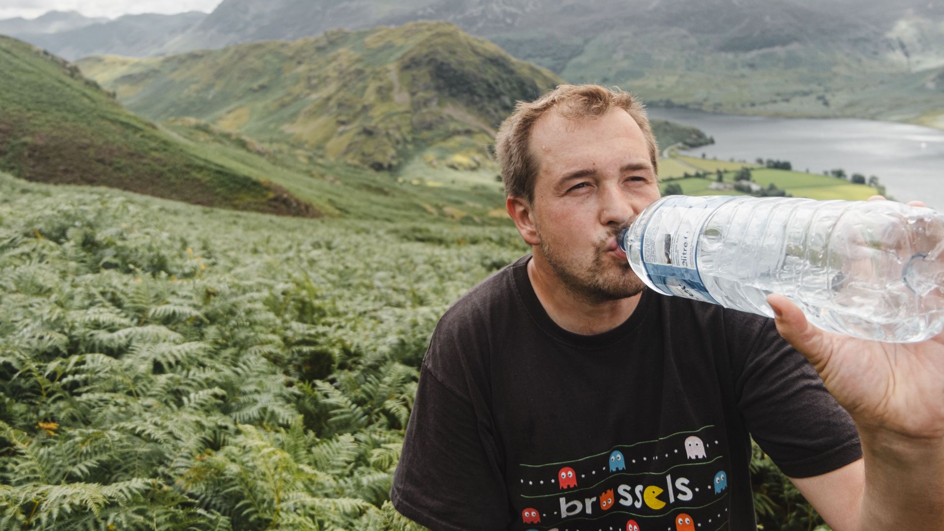 Adult man sipping water from bottle while resting near lush fern plants on slope of mountain on cloudy day in higlands