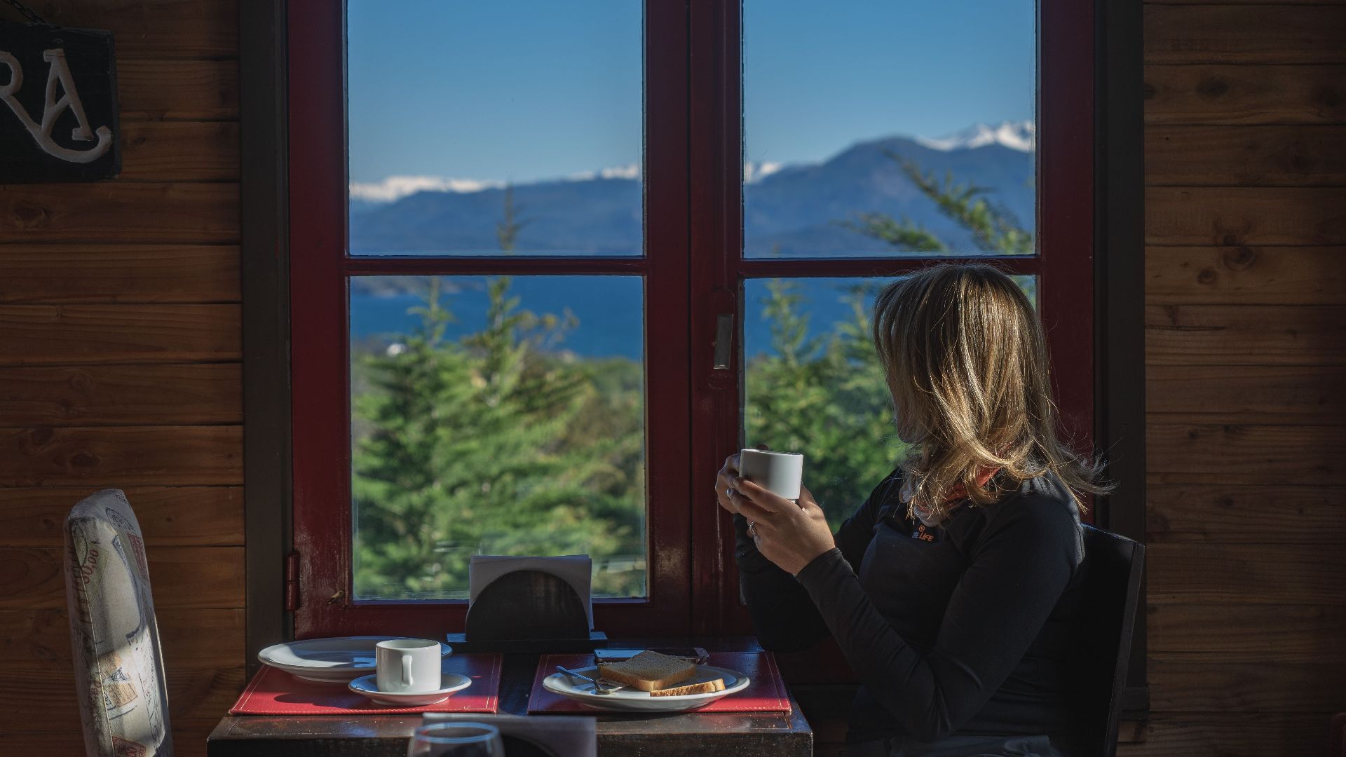 A woman enjoys coffee indoors with a scenic mountain view in Bariloche, Argentina.