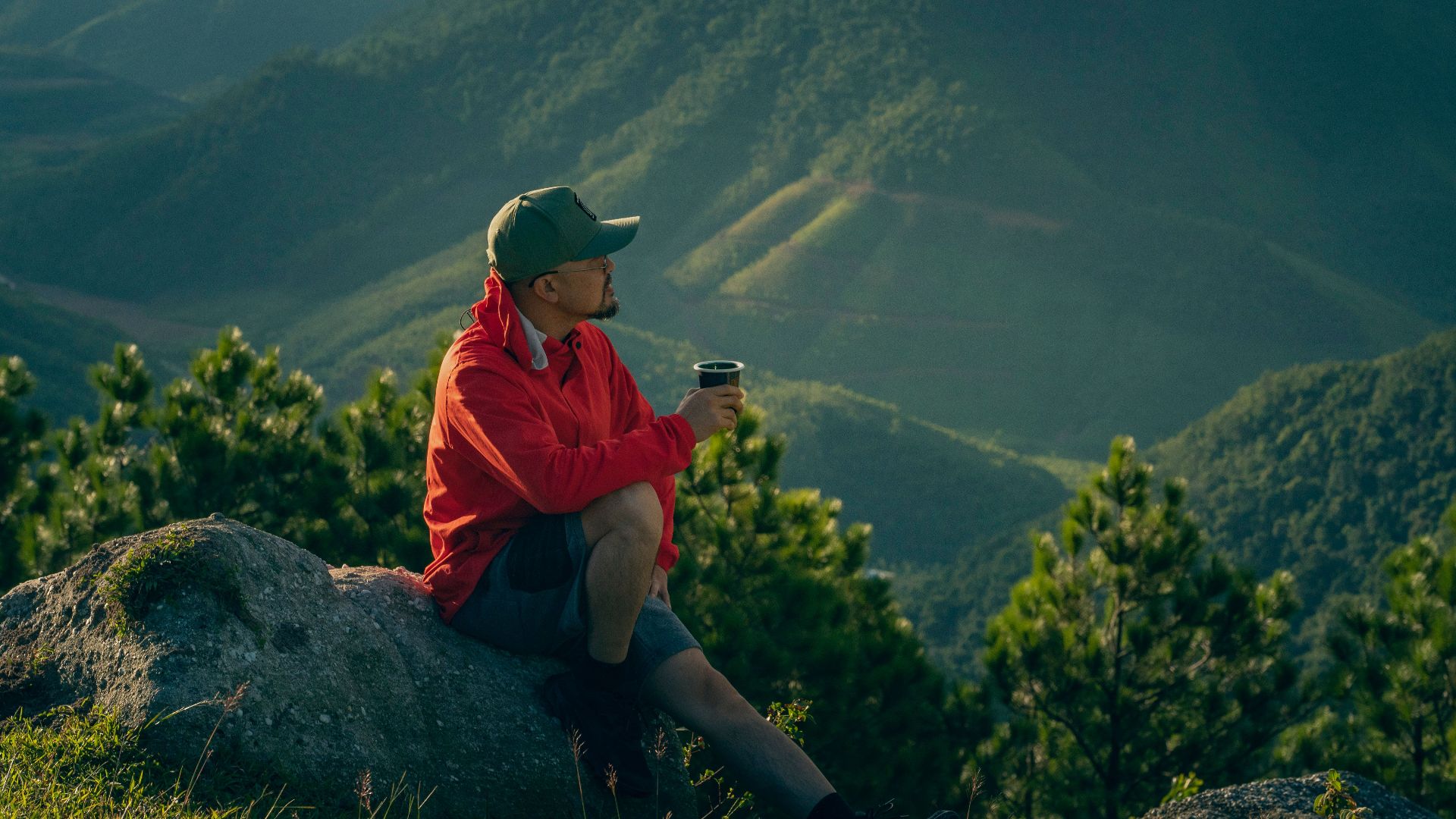 Man enjoys a serene moment in mountain scenery, sipping a drink at sunrise.
