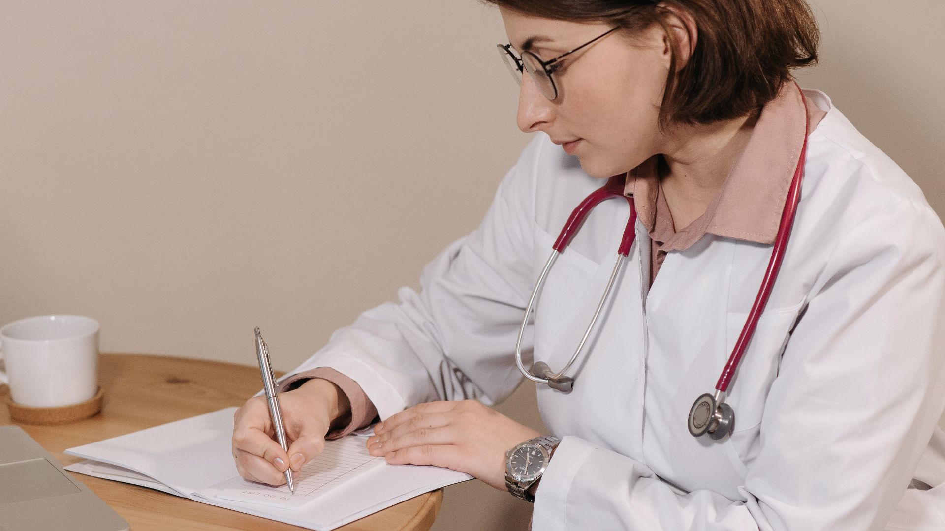 Woman doctor with stethoscope writing at desk with laptop and notes.