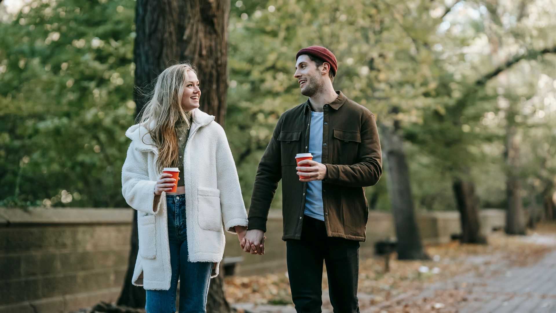 Happy stylish young man and woman in warm clothes holding hands and chatting while walking in autumn park and drinking takeaway coffee