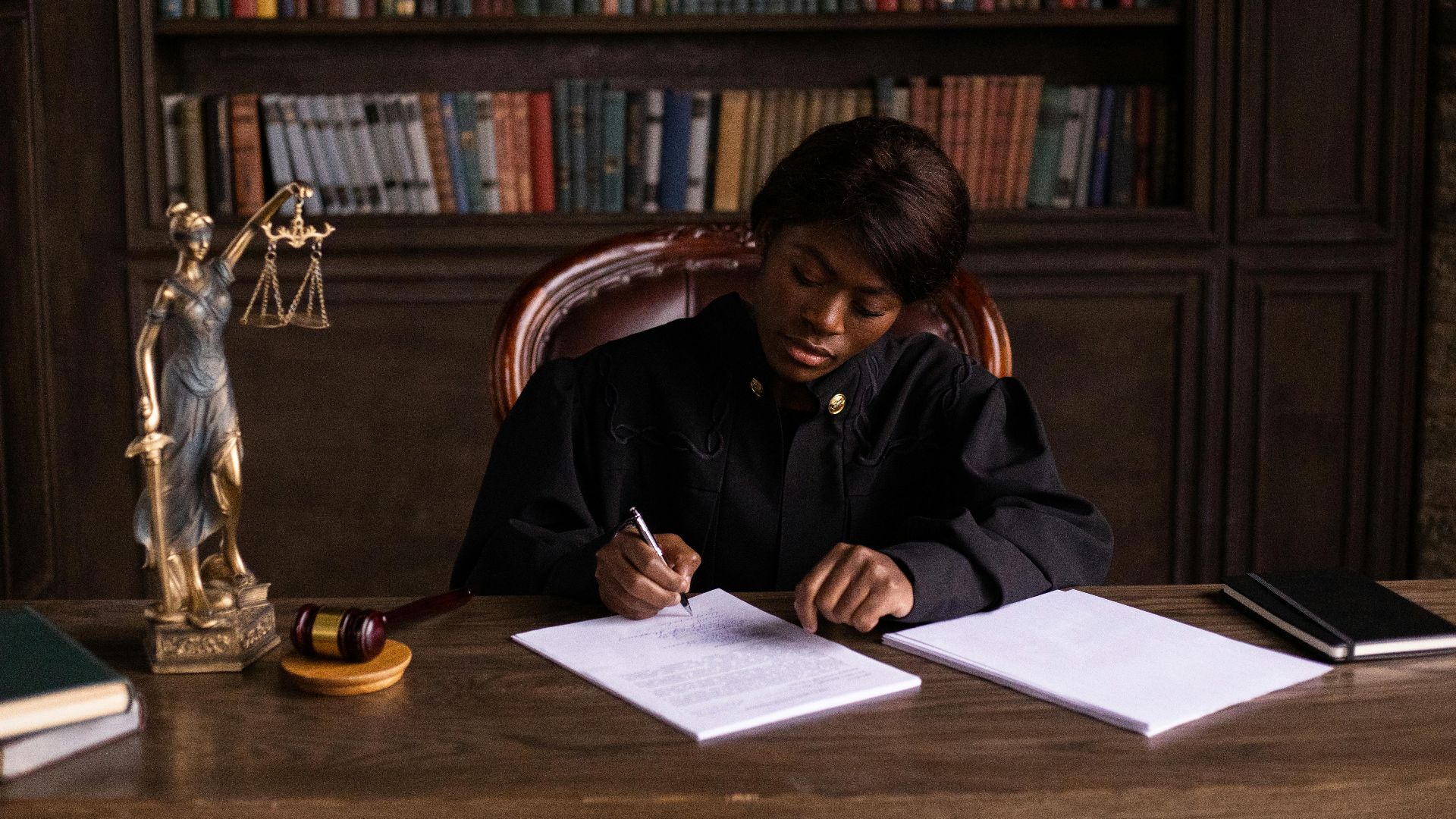 A judge in robes writing on a document at a desk in an office library with law books.