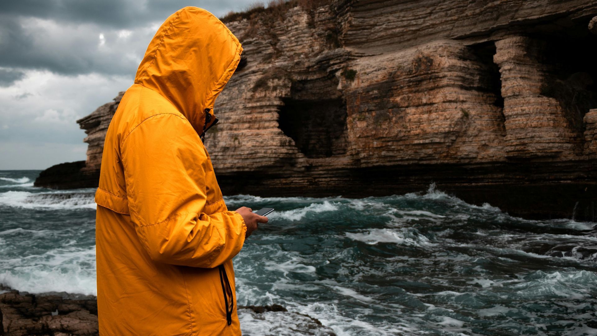 Person in a yellow jacket standing by rocky seashore texting during an overcast day.