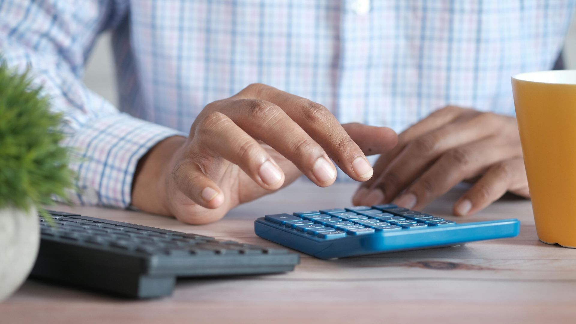 A person using a blue calculator on a wooden desk with a keyboard and plant nearby.