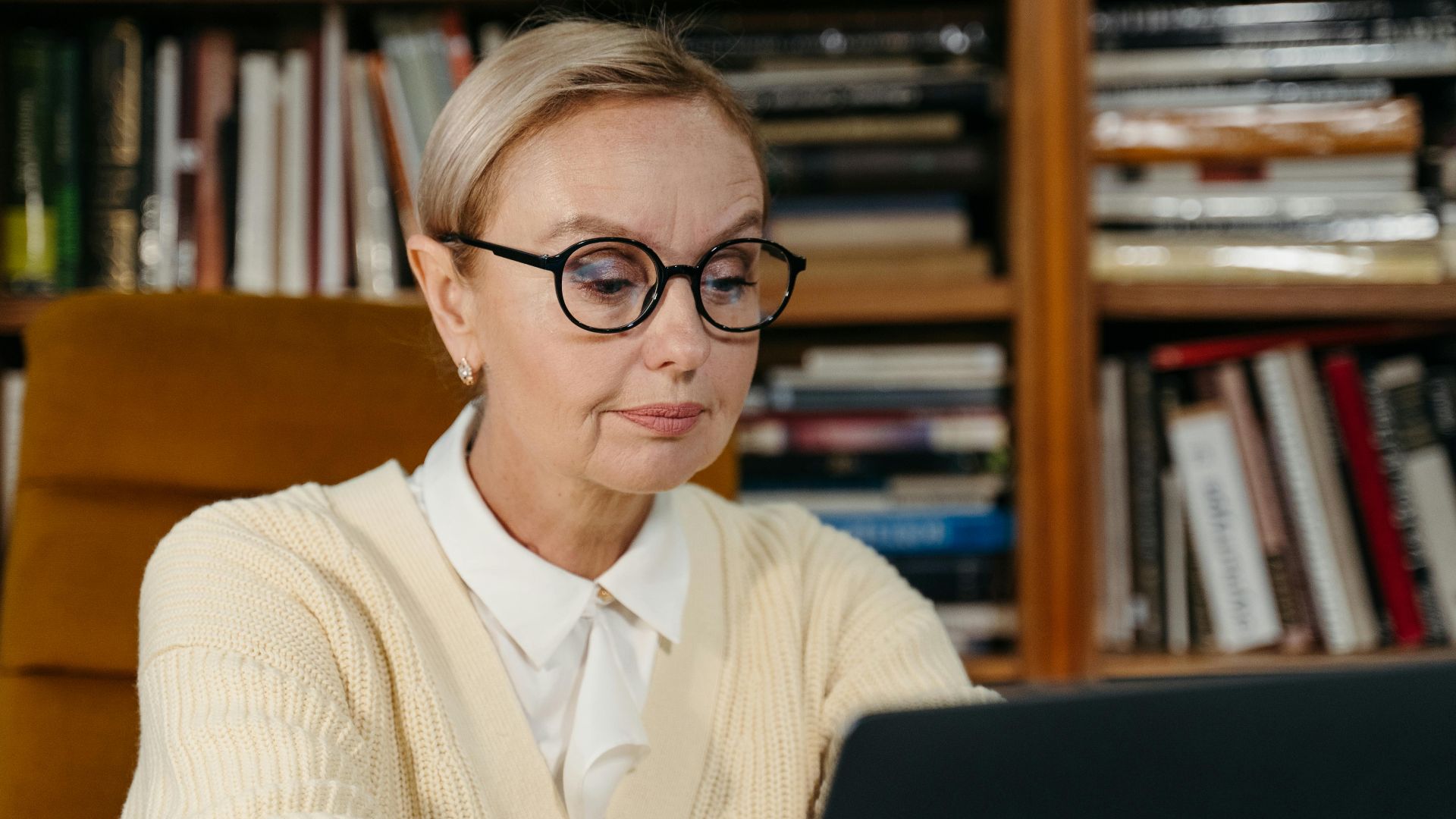 A senior woman wearing glasses focusing on her laptop in a cozy library environment.