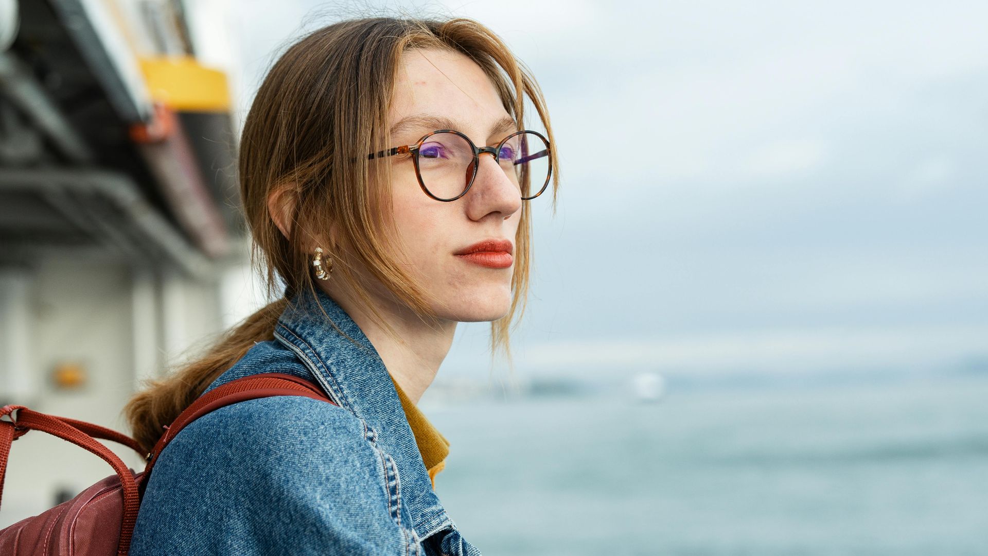 A woman in a denim jacket enjoys the sea view from a sailboat, leaning on the railing.
