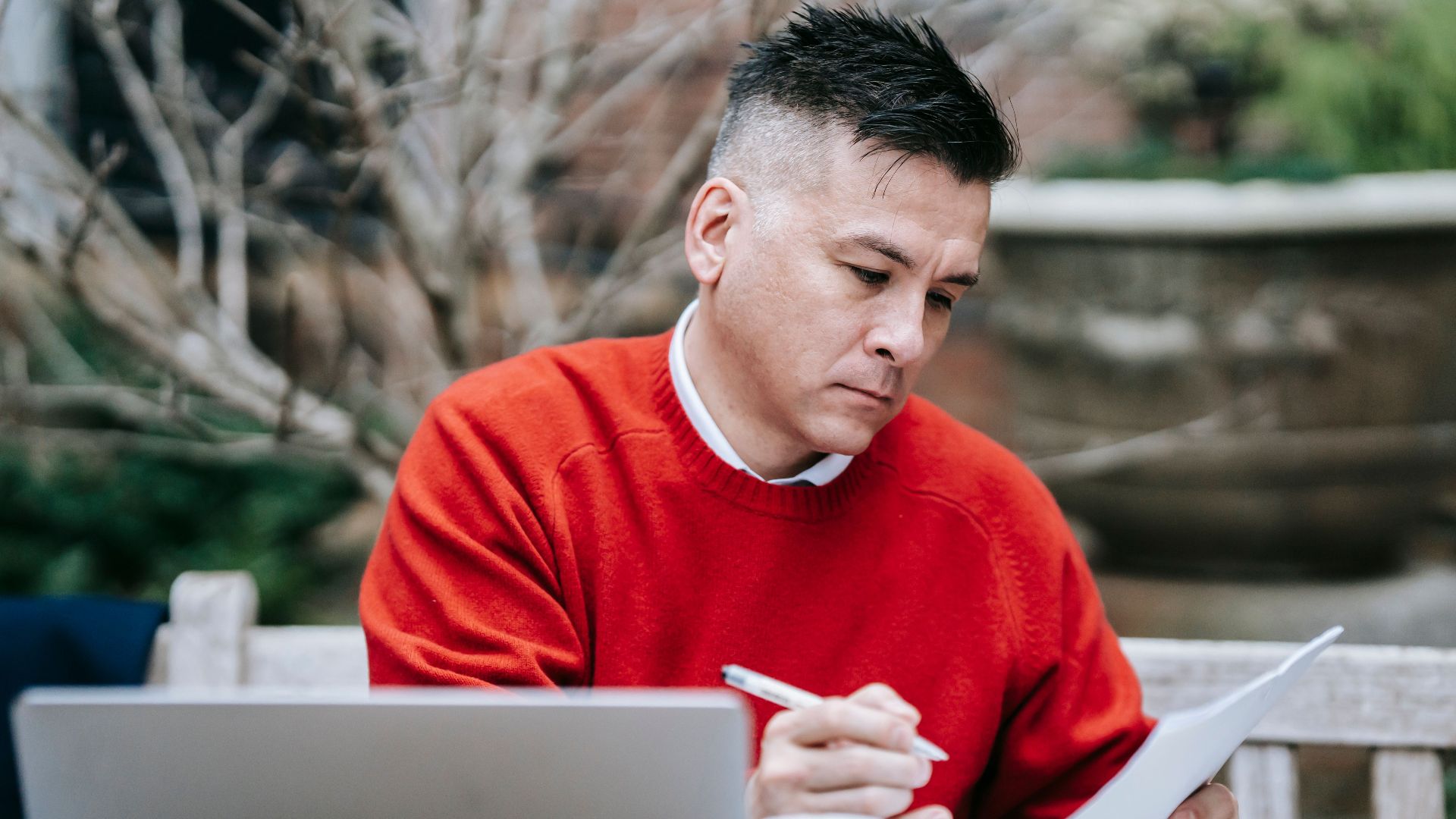 A man in a red sweater concentrating on paperwork while working remotely with a laptop outdoors.