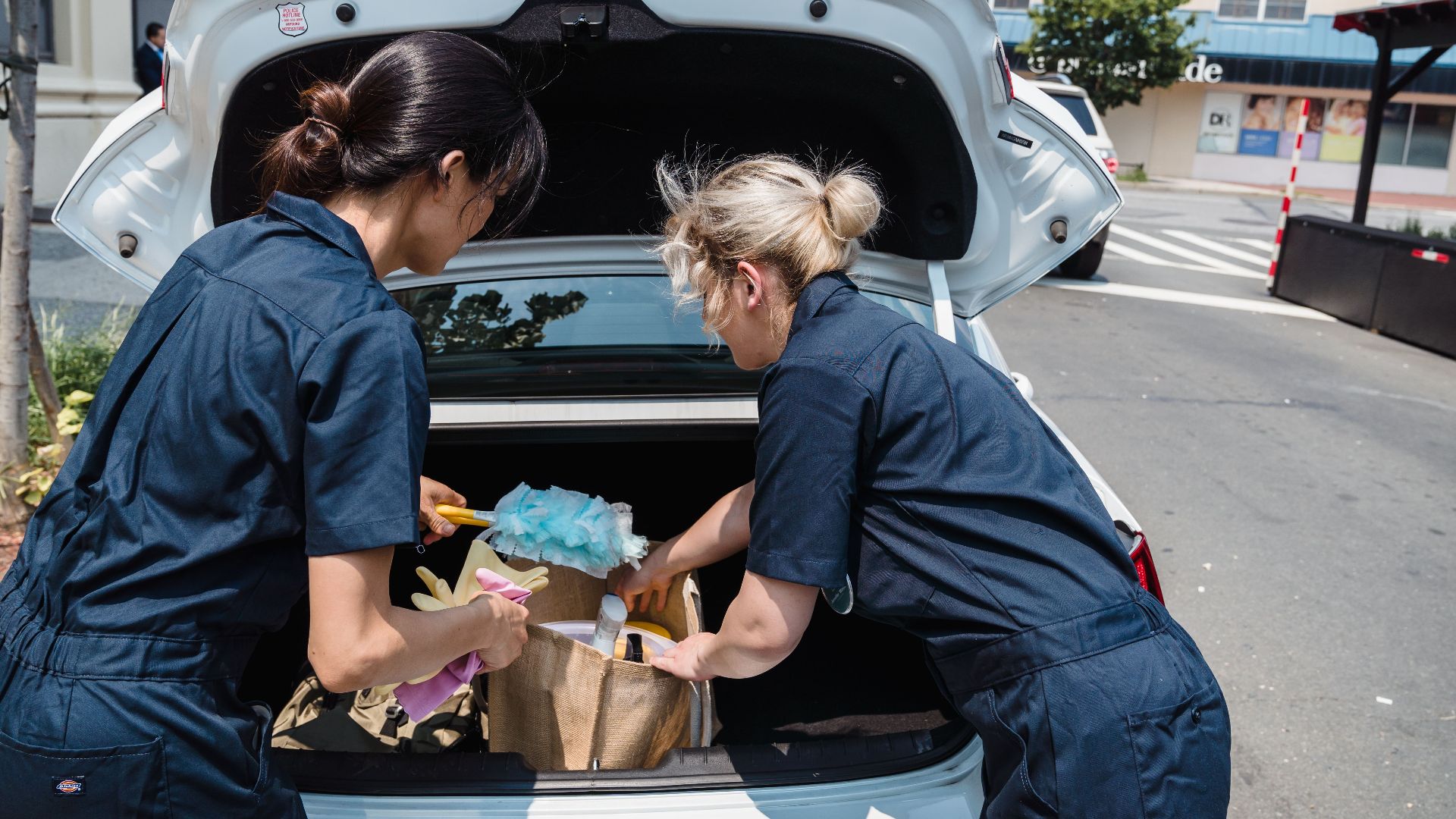 Two women in uniforms unloading cleaning supplies from a car trunk in an urban area.