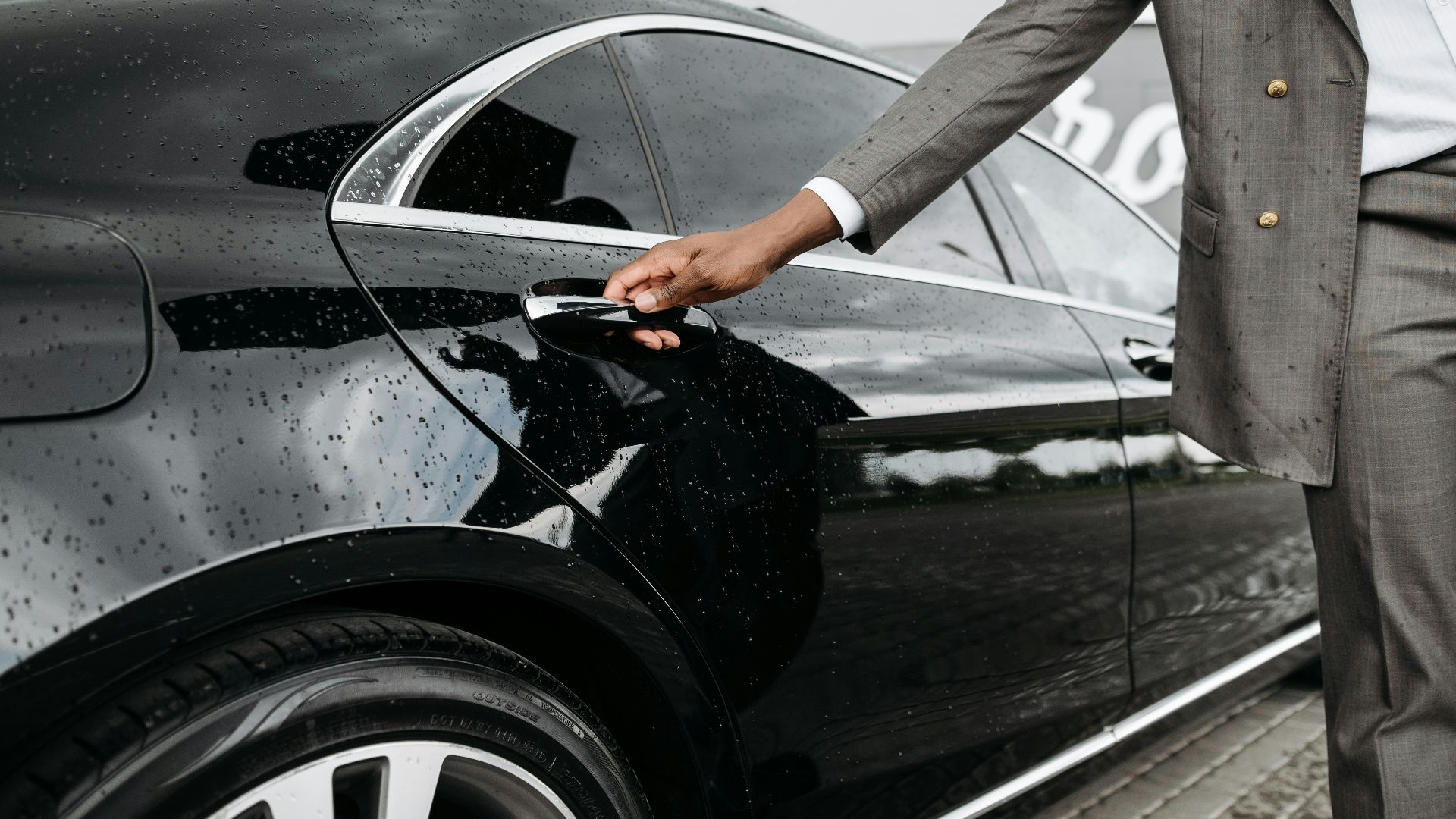 A man in a suit opens a luxury car door under raindrops on stone pavement.
