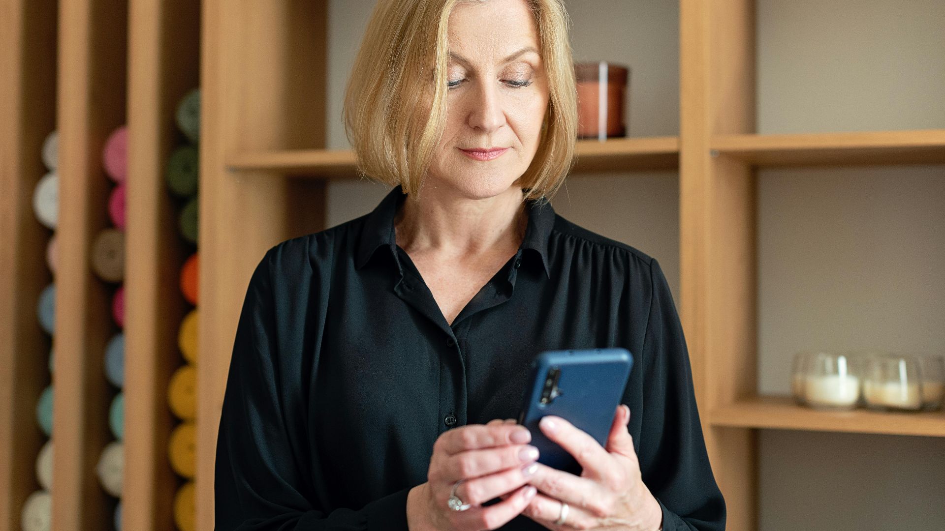 Professional woman in black attire using a smartphone indoors with shelving background.