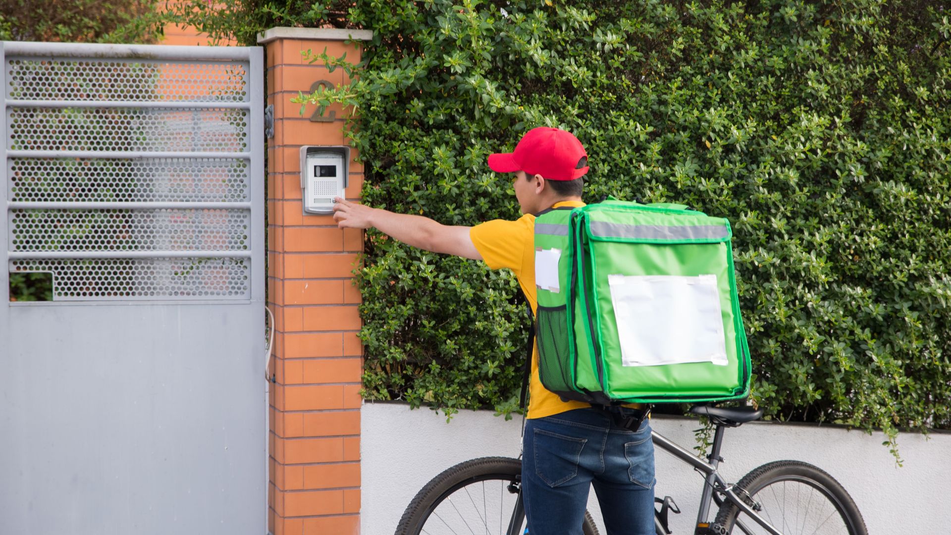 Courier in yellow shirt and red cap delivering a package on a bicycle, pressing a doorbell outdoors.