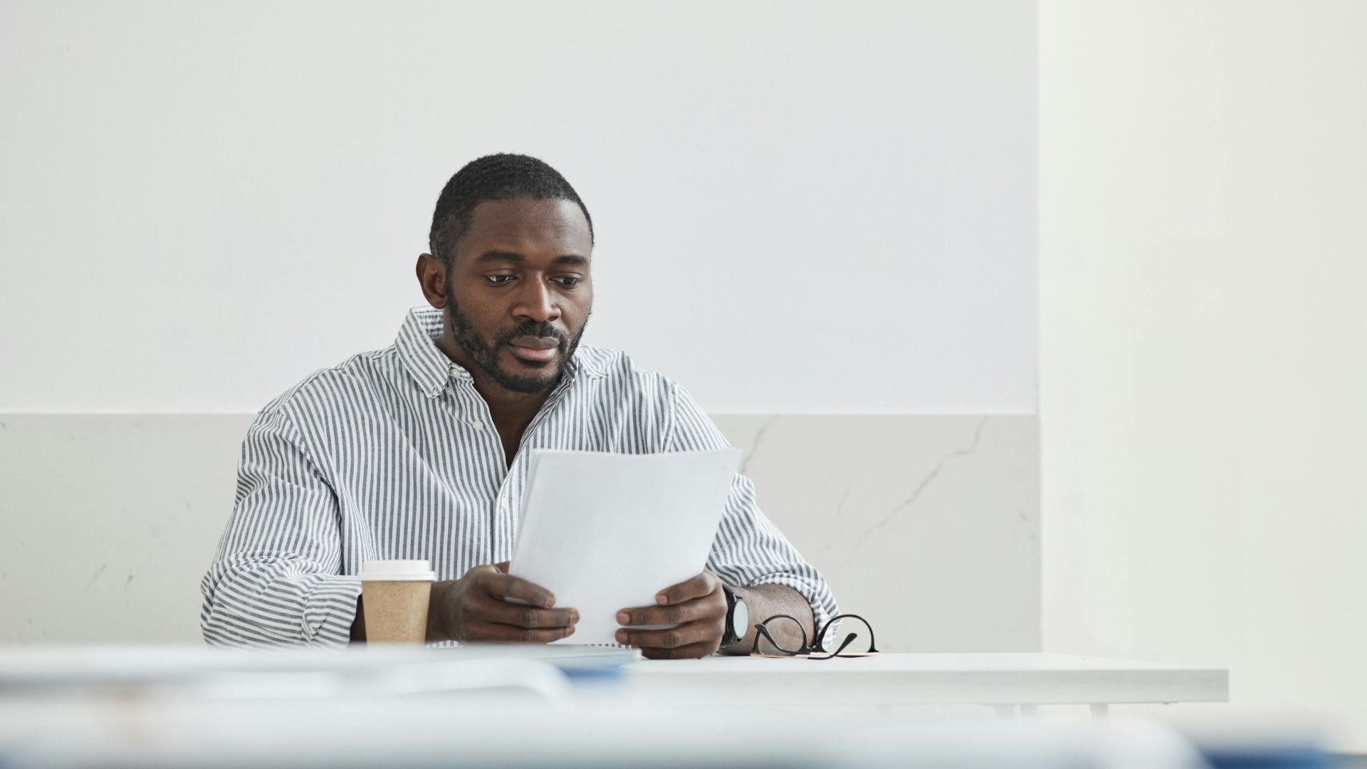 African American man sitting indoors, reading papers with a coffee cup nearby.