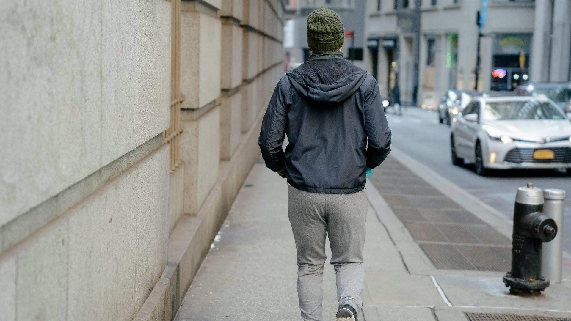 Back view of anonymous male in gray windbreak jacket and sweatpants and knitted green hat running on paved sidewalk in street of city