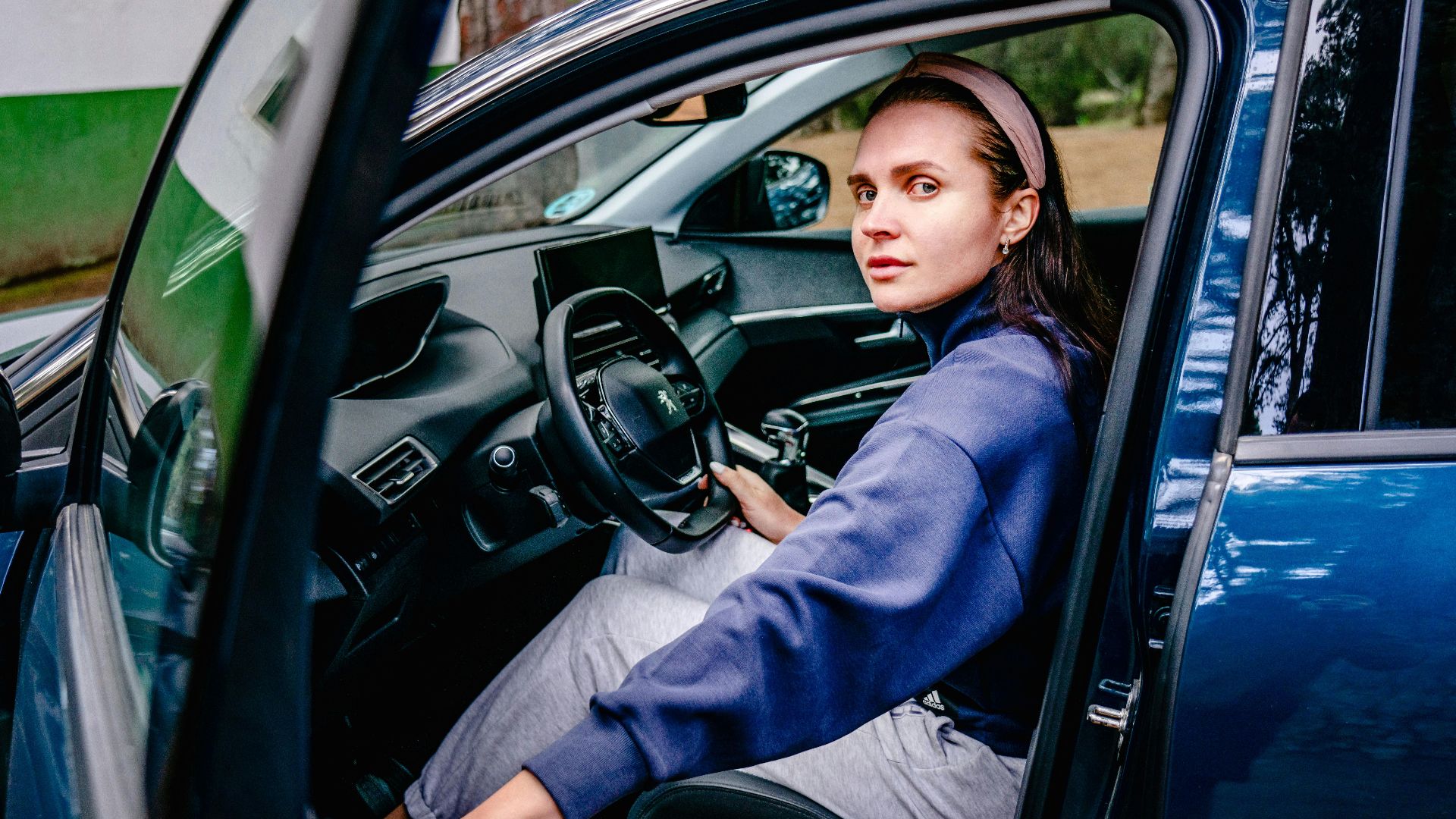 Woman sitting in the driver's seat of a car with an open door, looking outside. Natural setting.