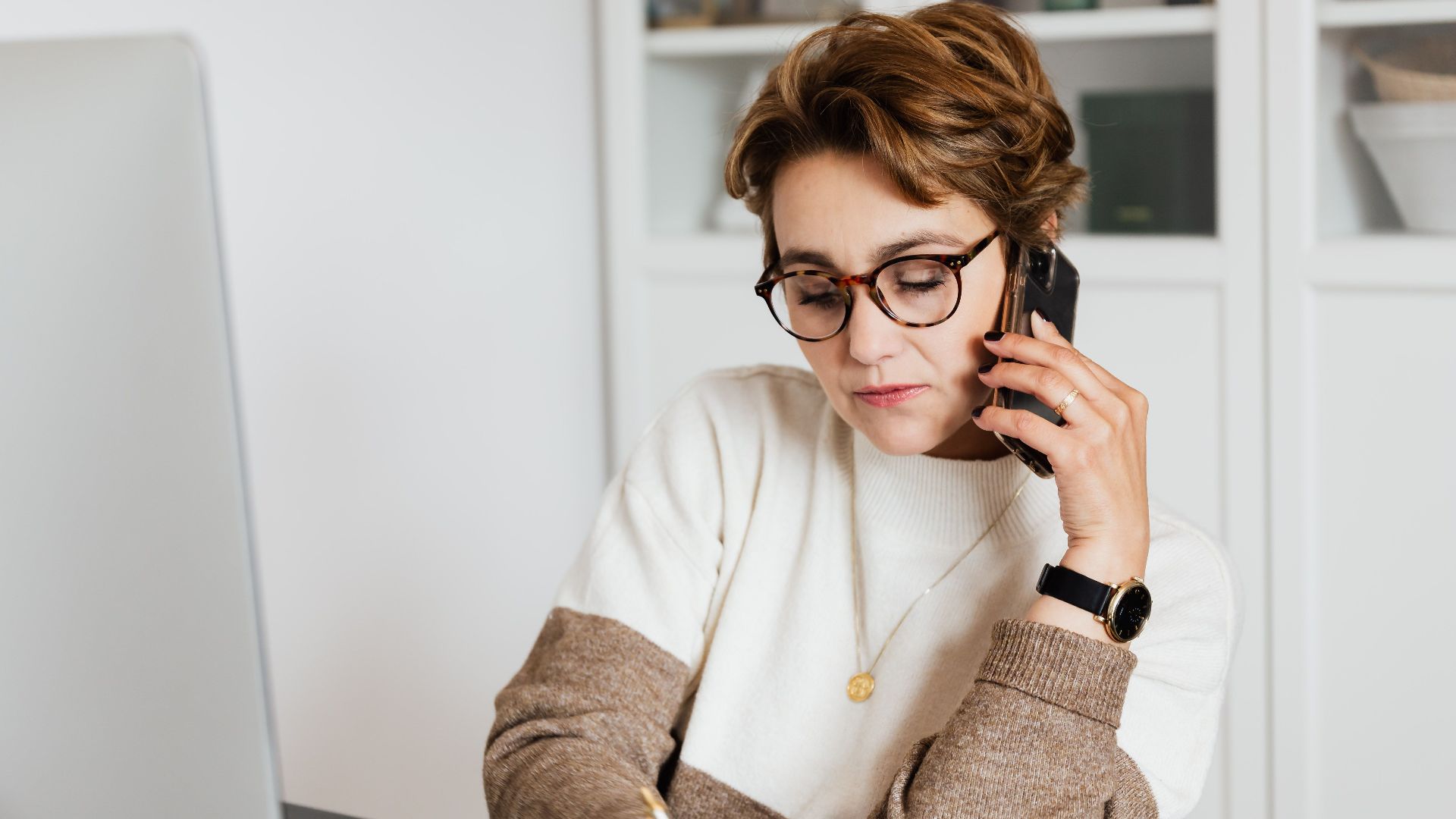 Good looking short haired female worker listening to work instructions on mobile and noting down information in modern office