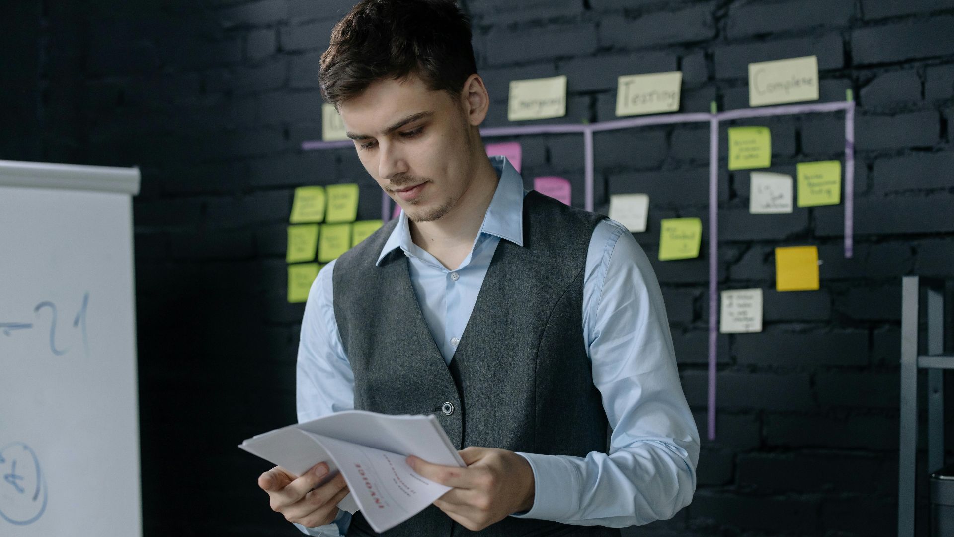 A young man reviewing documents in an office with a black brick wall and sticky notes background.