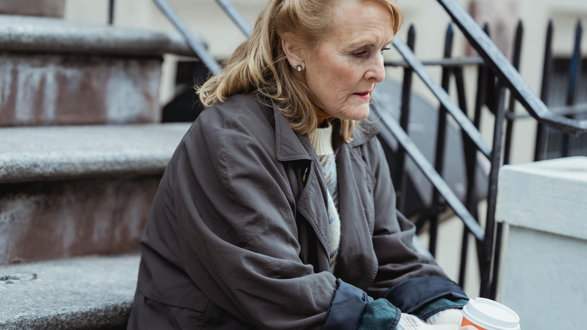 Reflective senior female in outerwear with hot drink to go looking down while sitting on city stairs