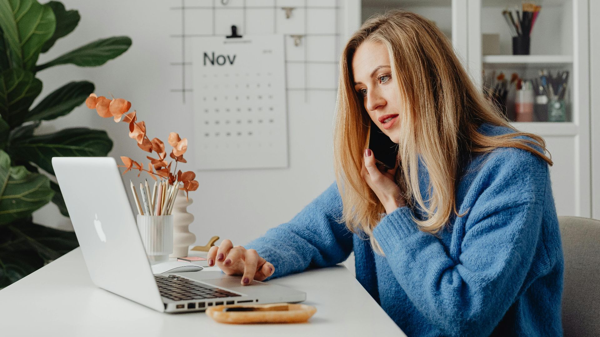 Woman multitasking with laptop and phone in a home office setting.