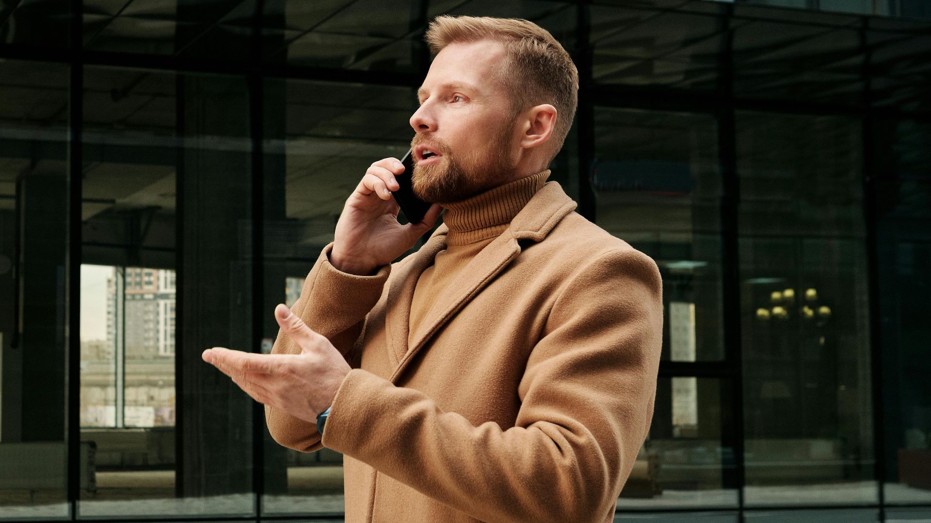 Confident businessman in a brown coat talks on phone outdoors in city.
