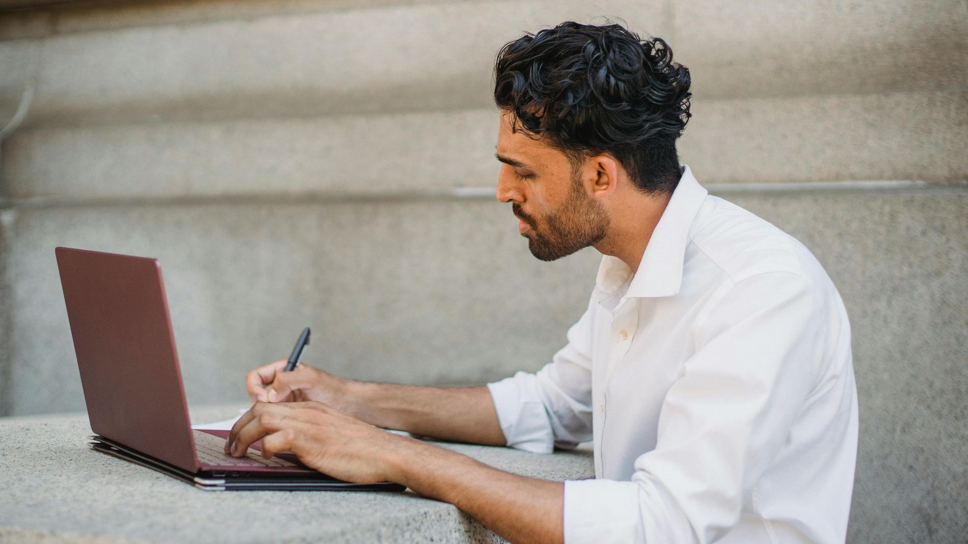 A man writing on a laptop outdoors, seated on a stone surface and focused on work.