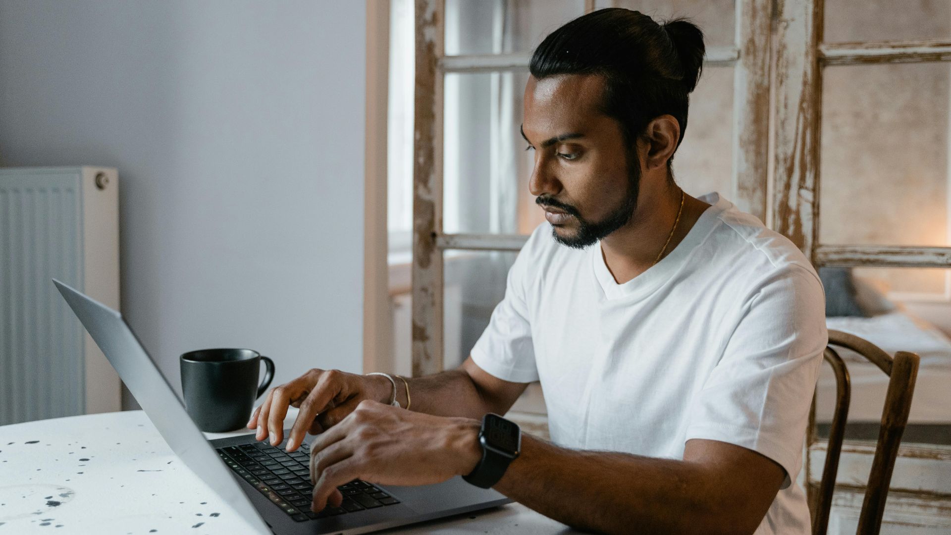A bearded man is focused while typing on his laptop at home, seated at a desk.