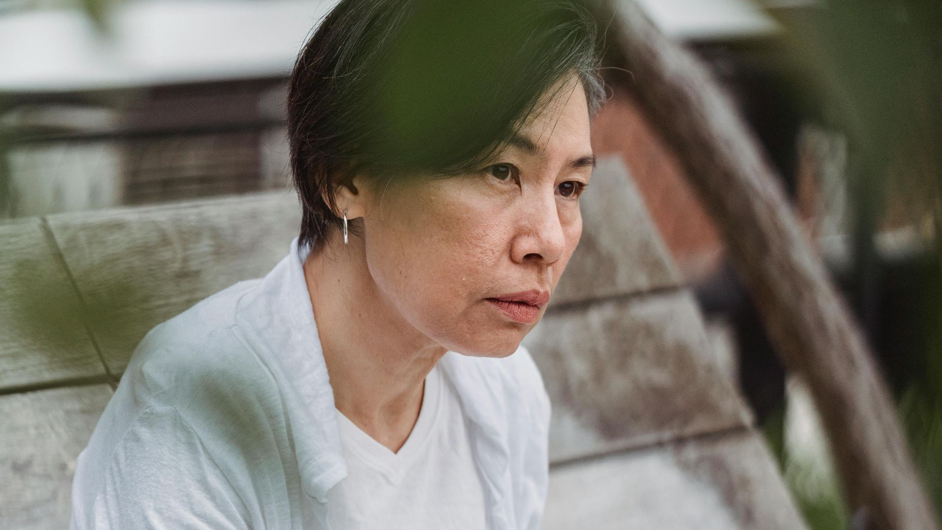A thoughtful woman sketches in a notebook while sitting on a wooden bench outdoors.