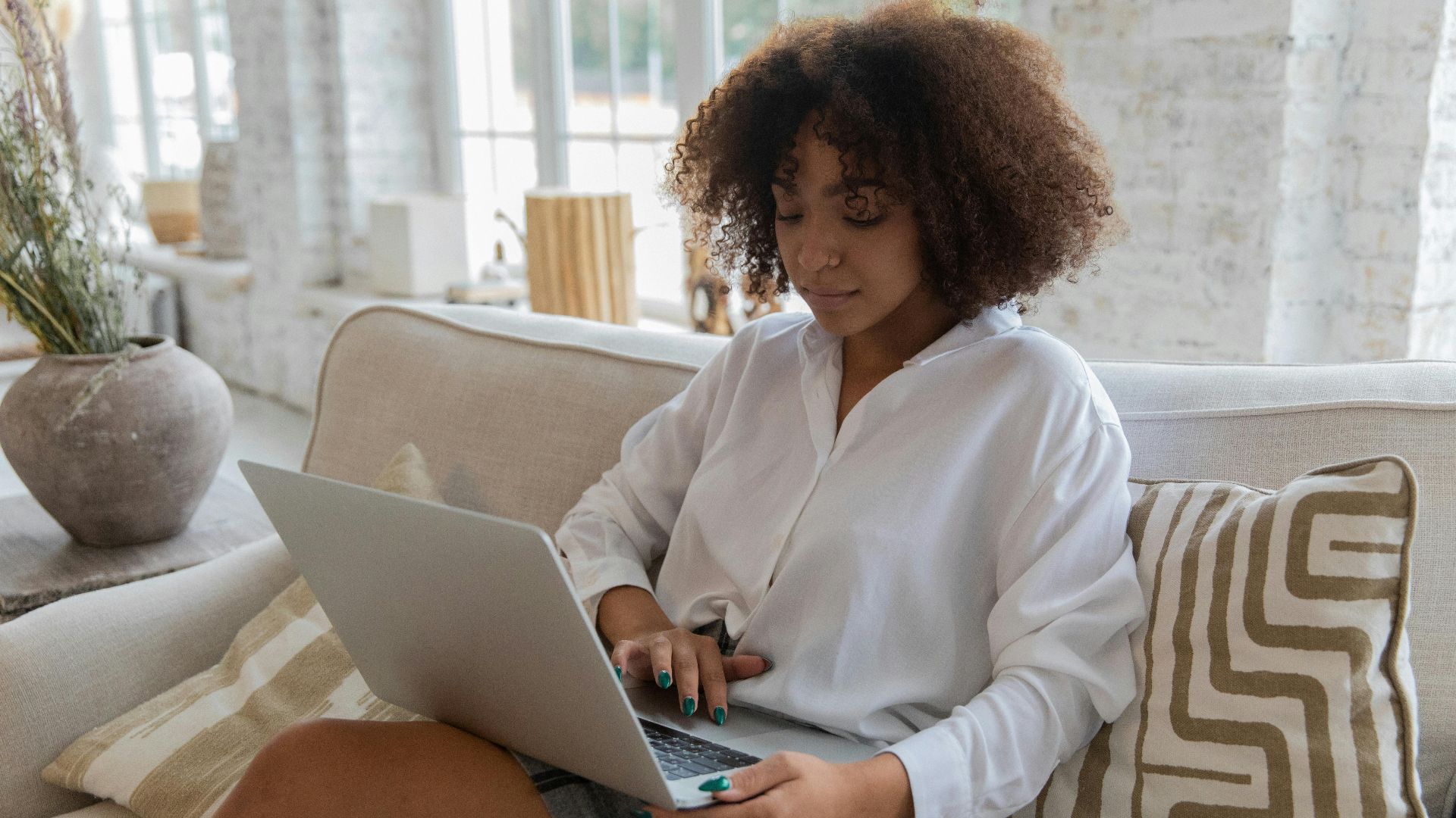 Concentrated young African American female student with curly hair in casual clothes browsing laptop while sitting on sofa during online lesson at home