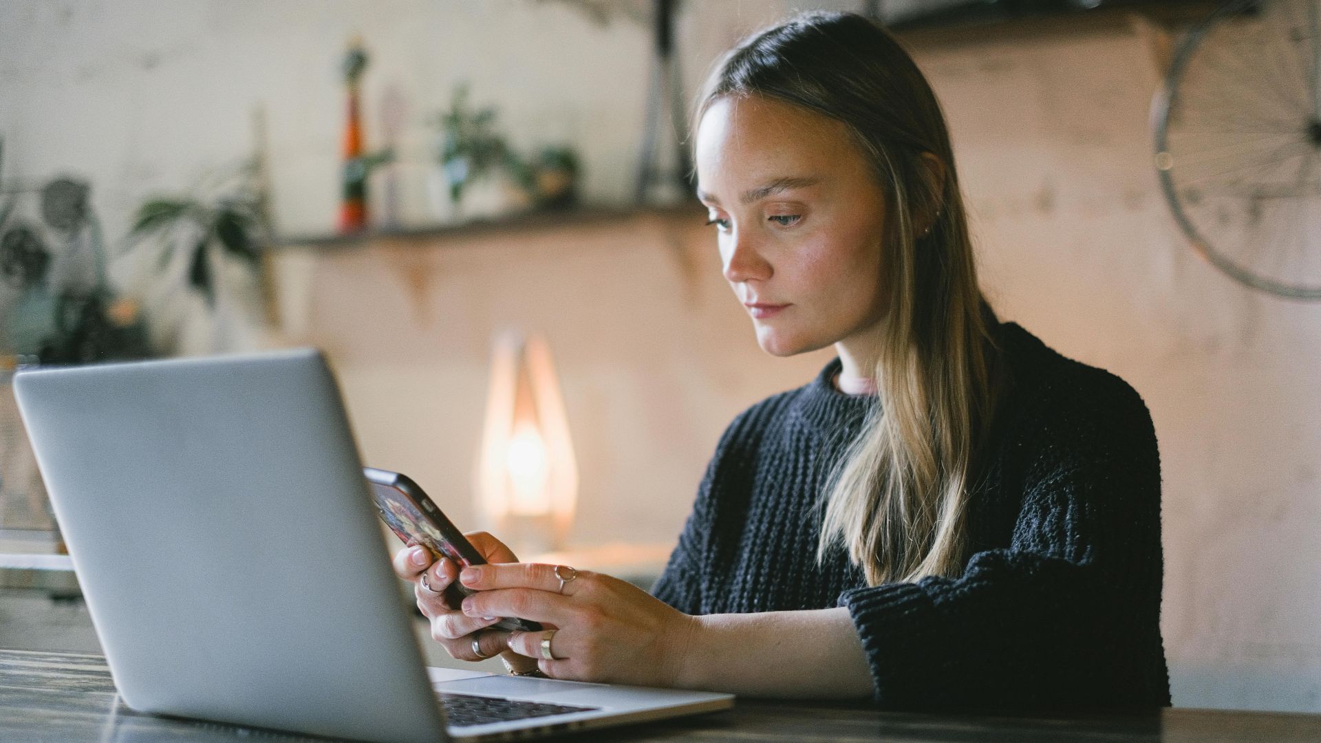 Concentrated female freelancer text messaging on cellphone while sitting at table with netbook during online work on blurred background
