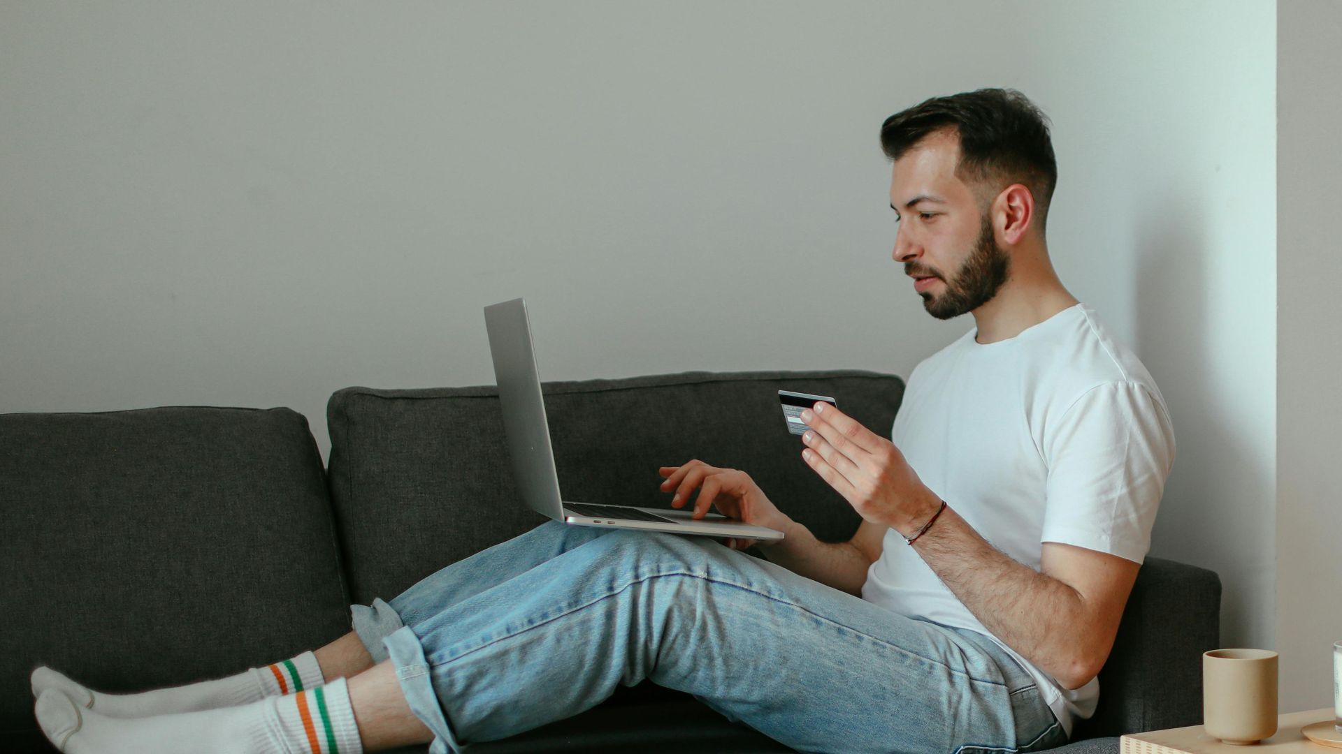 Adult man using laptop for online shopping on sofa, holding credit card.