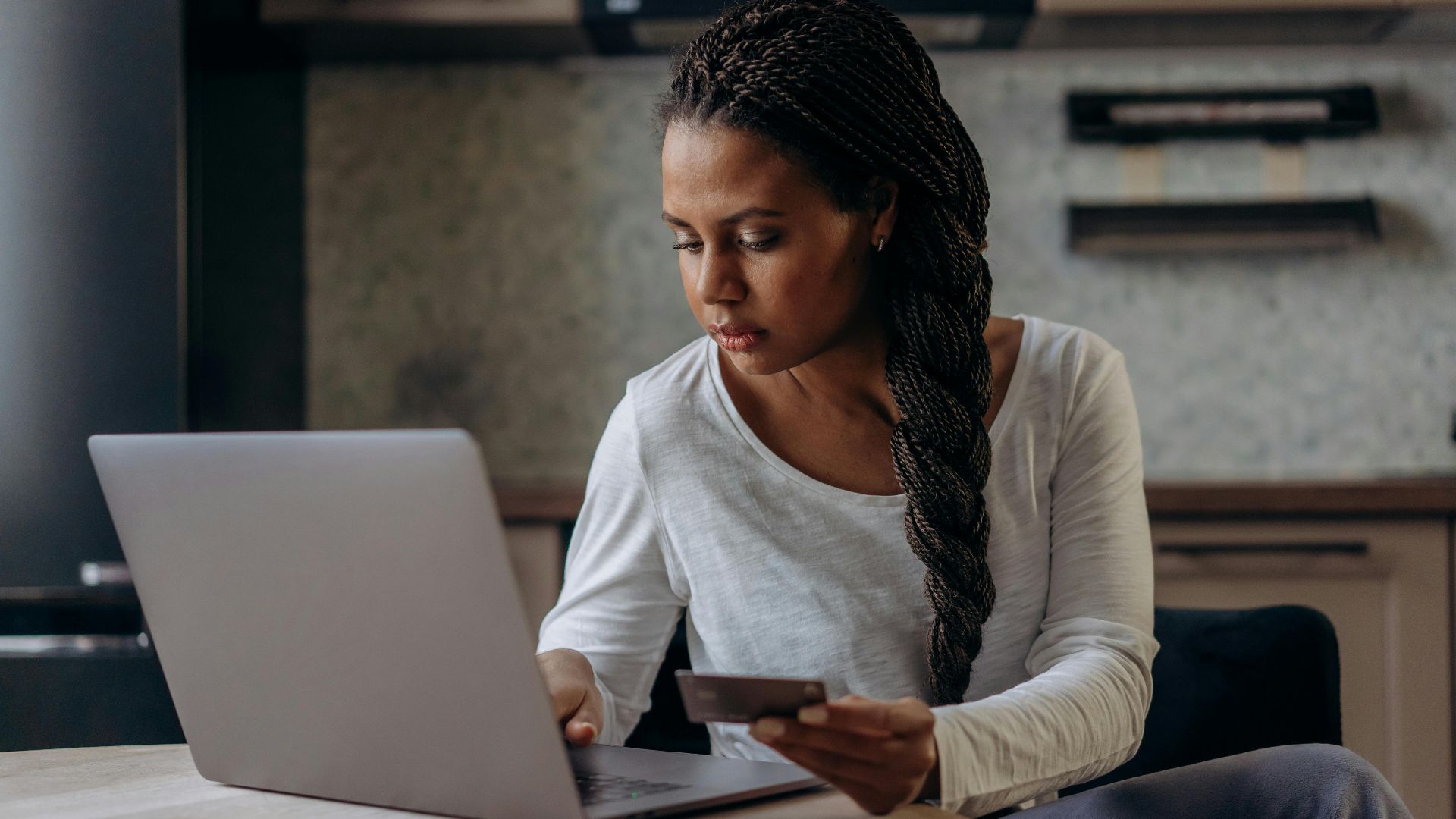 A woman with braided hair uses her laptop and credit card for online shopping indoors.
