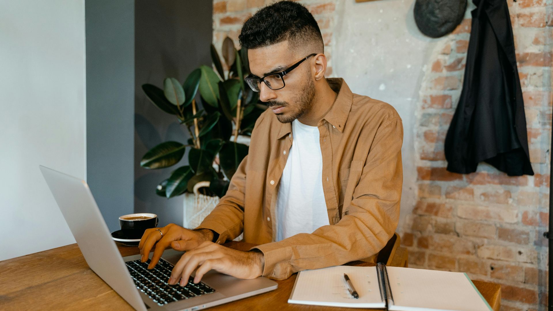 Man in casual office setting typing on laptop with coffee and notebook on desk.