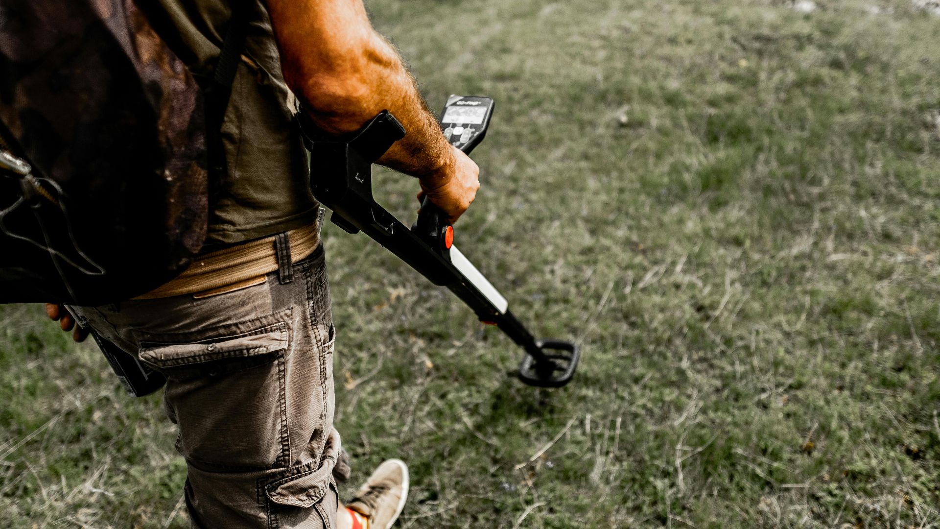 Person using a metal detector on a grassy field wearing casual attire.