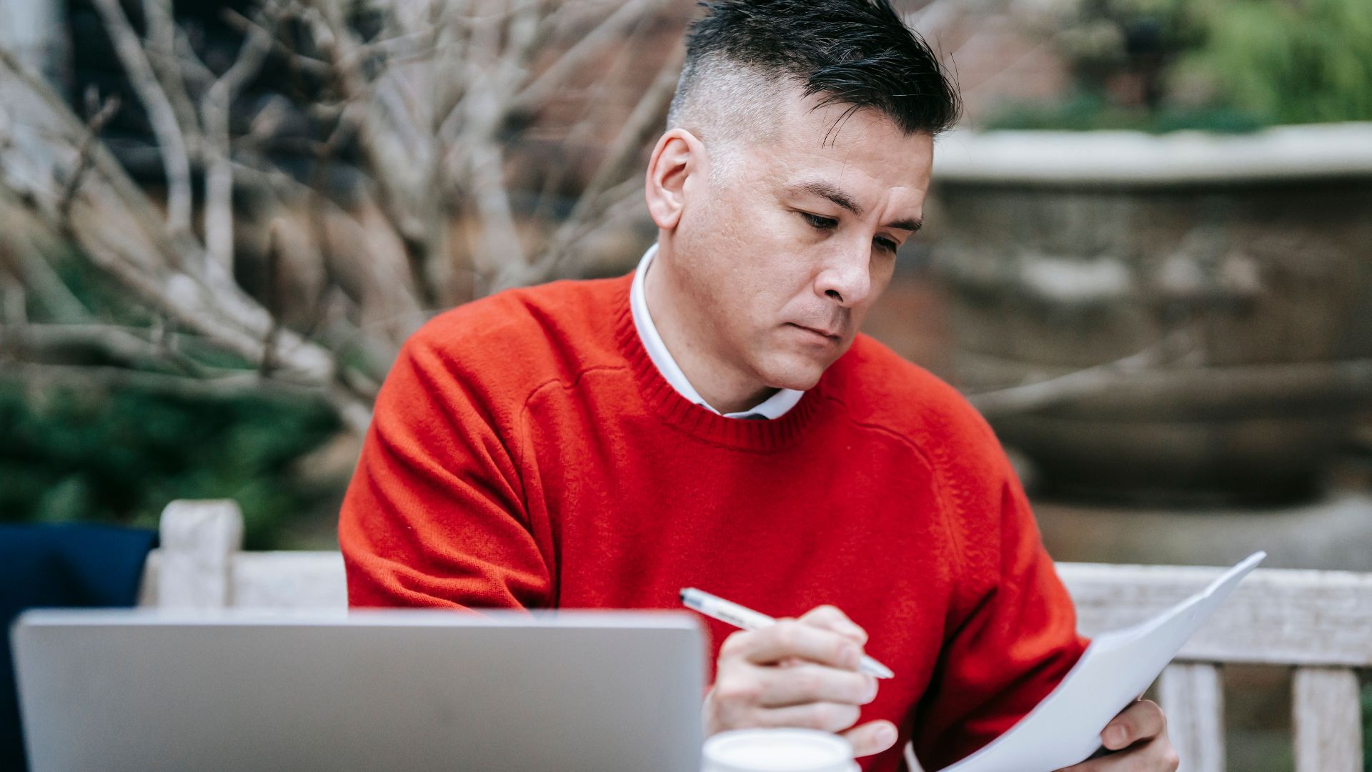 A man in a red sweater concentrating on paperwork while working remotely with a laptop outdoors.