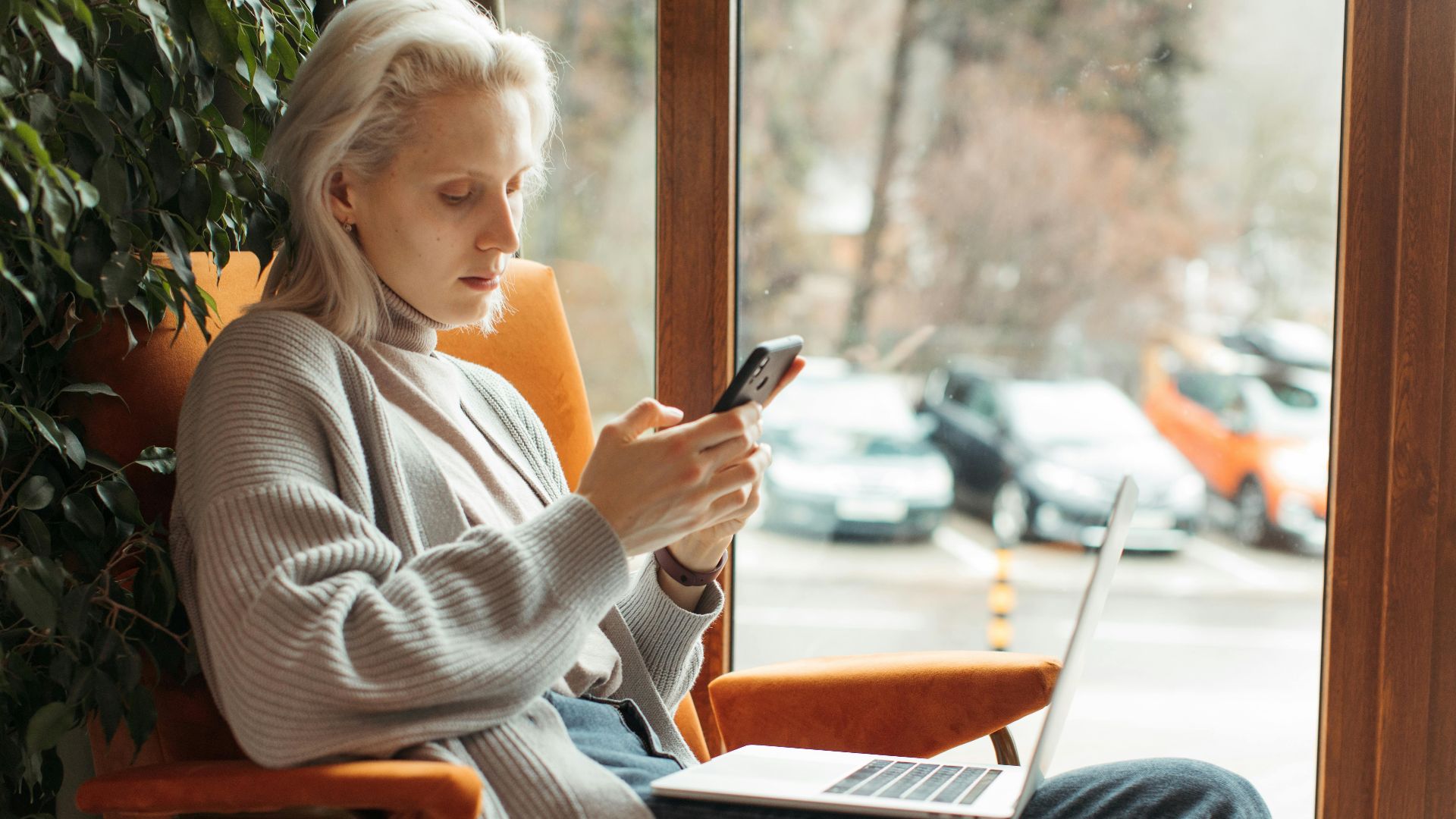 Woman sitting by a window, focused on her smartphone and laptop, in a cozy setting.