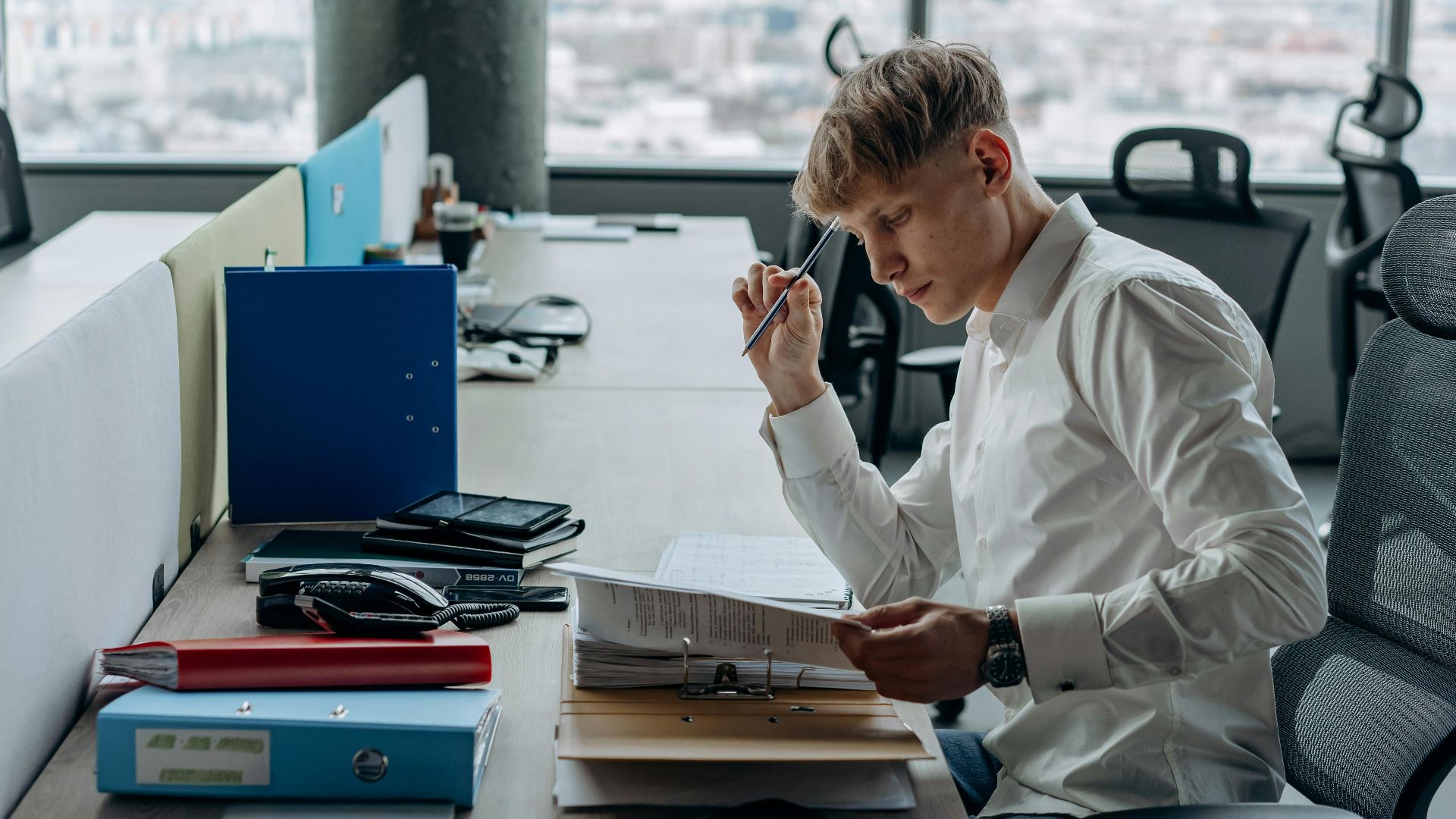 Man in white shirt concentrating on paperwork at modern office desk with city view.