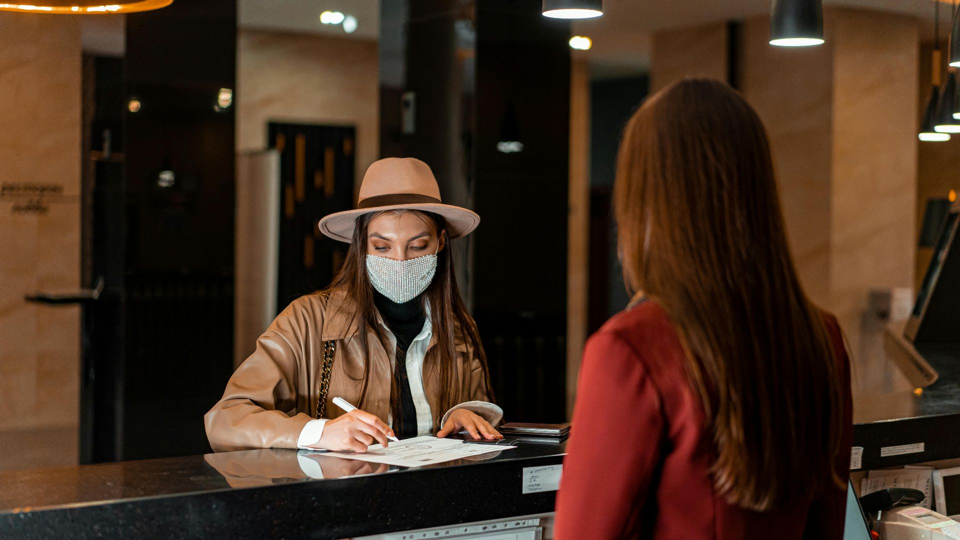 A woman in a hat and face mask checks in at a hotel reception, interacting with a receptionist.
