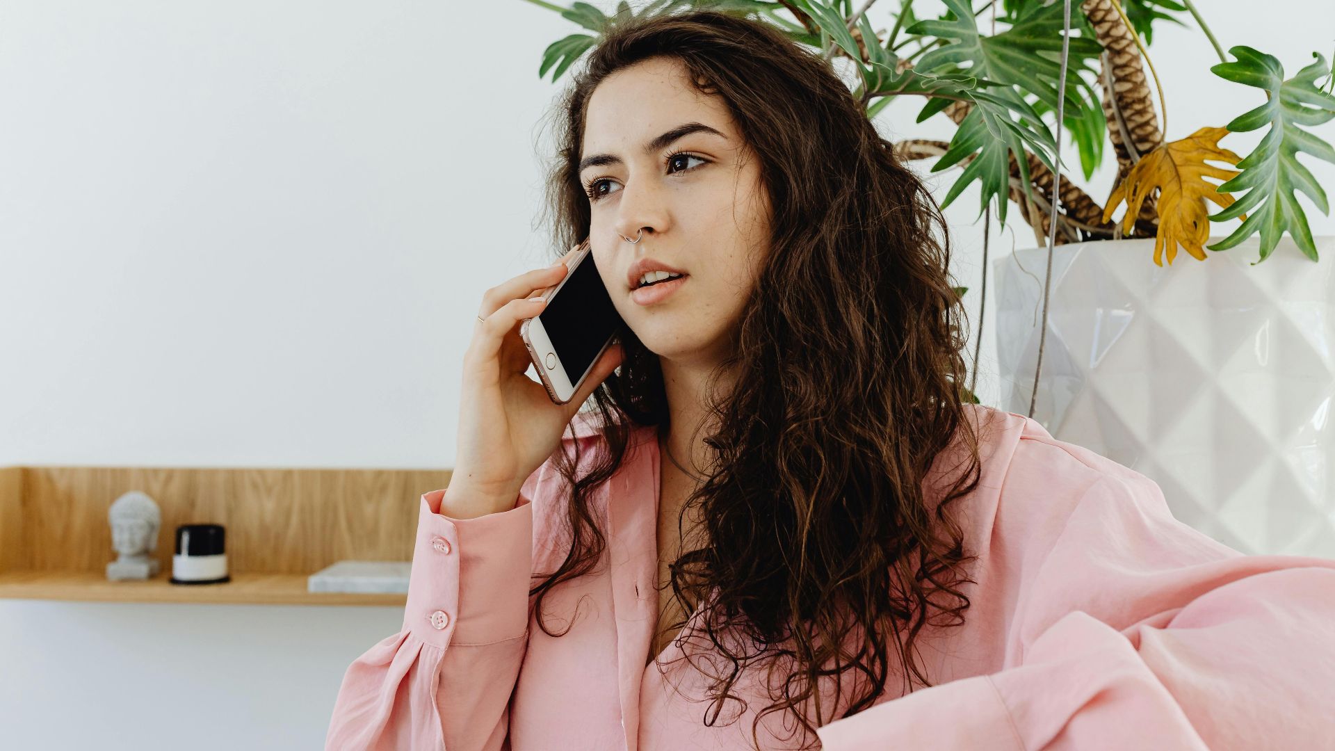 Confident young woman talking on smartphone in a modern interior with green plant background.